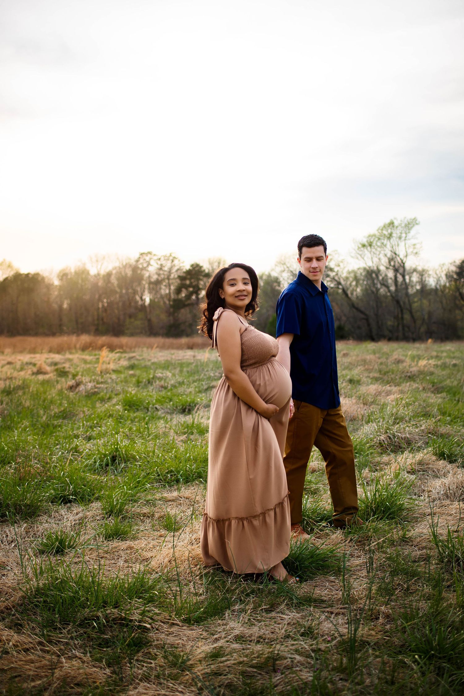 A couple poses in a sunlit field during sunset, with the expectant mother wearing a flowing tan dress with raleigh maternity photographer