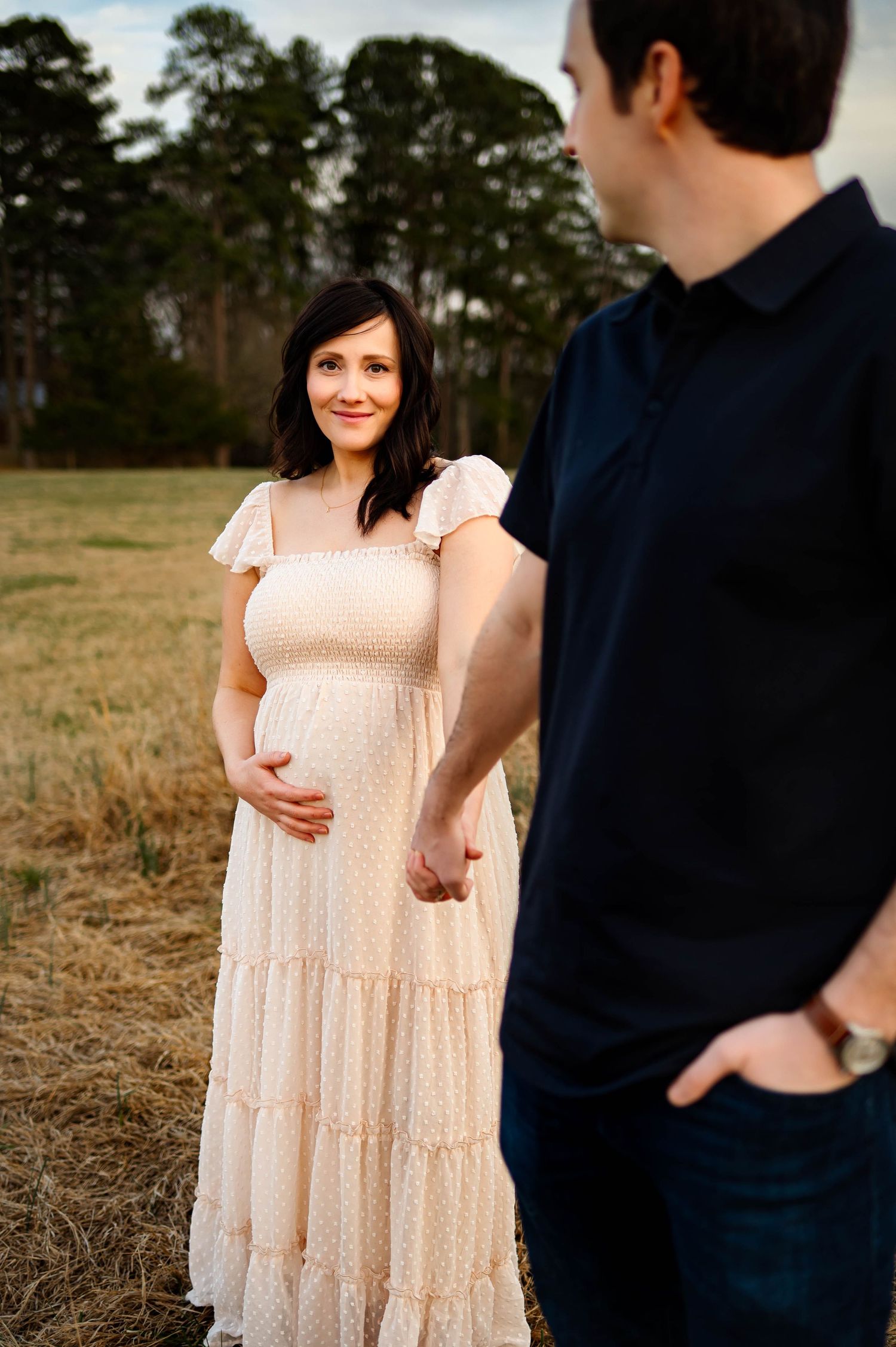 A couple poses in a field at sunset, with her wearing a flowing tiered maxi dress while expecting with the best cary maternity photographer