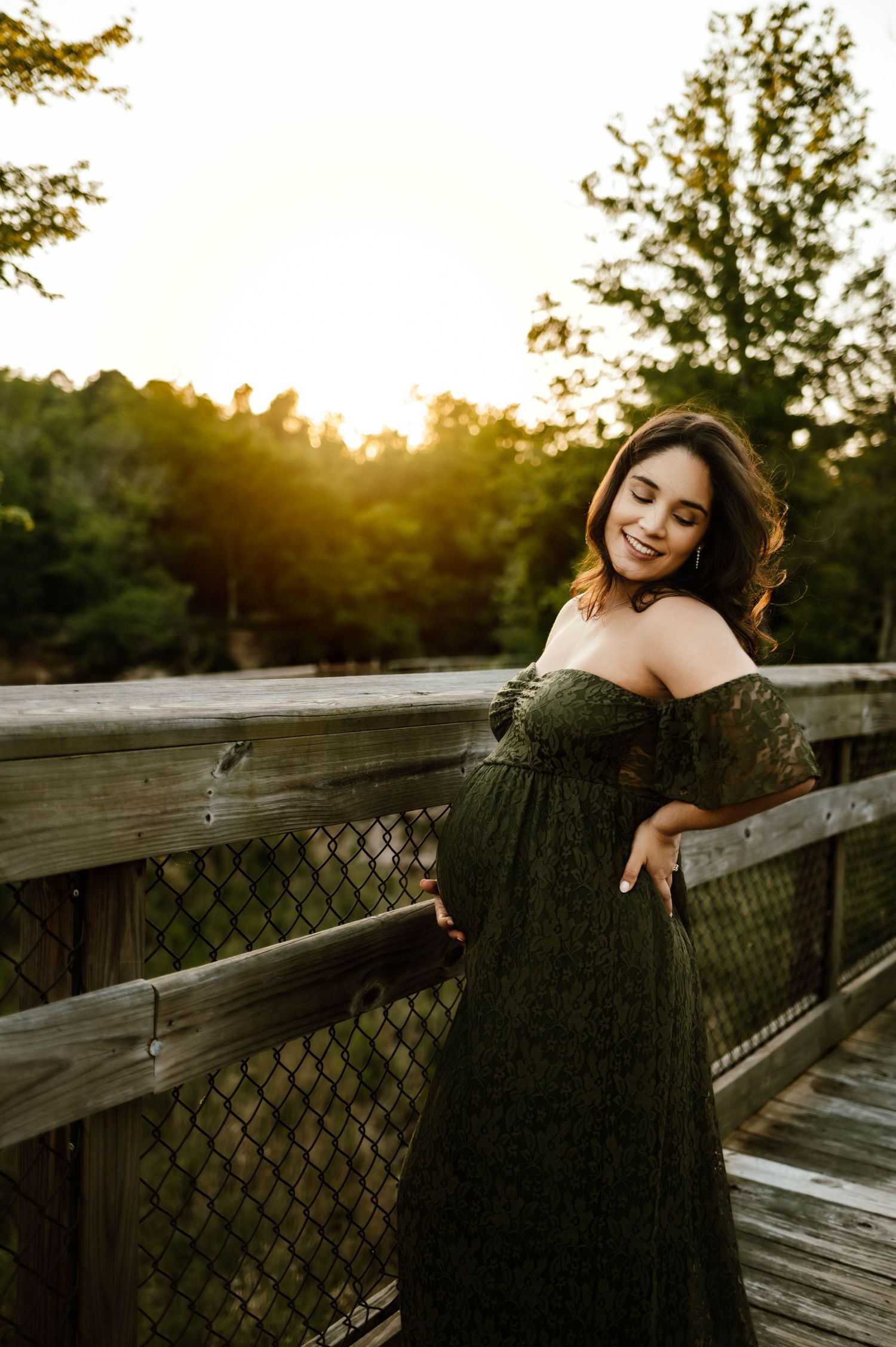 Golden sunset illuminates a wooden boardwalk where mother poses in an olive green off-shoulder dress for raleigh maternity photographer