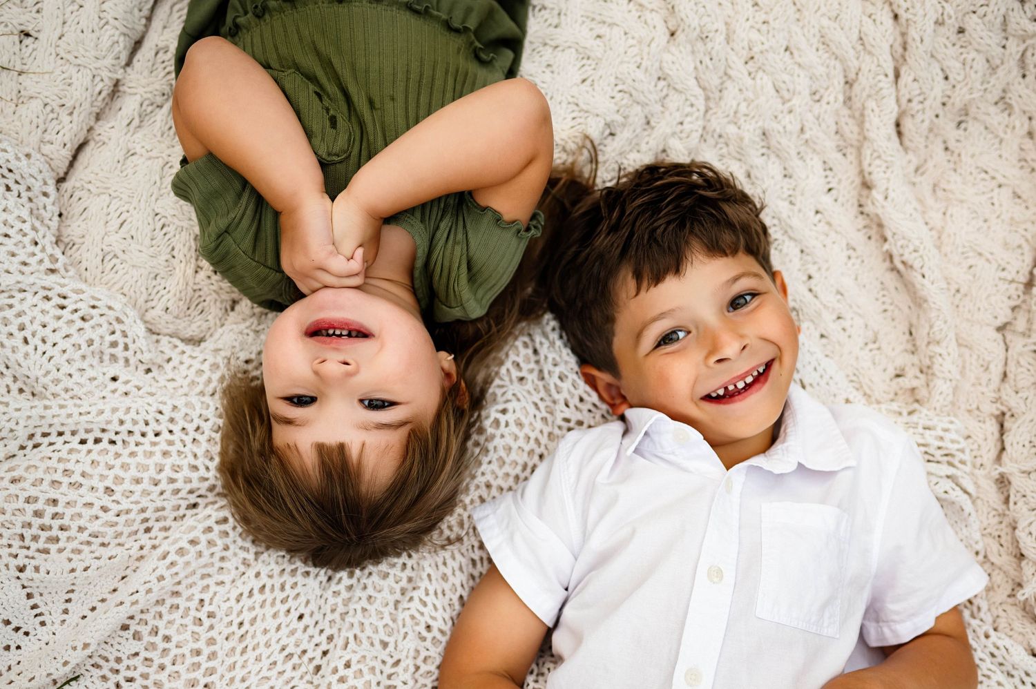 Two young children lying on a beige blanket while smiling at the camera for a photograph in Wake Forest  NC with Emma Branch Photography