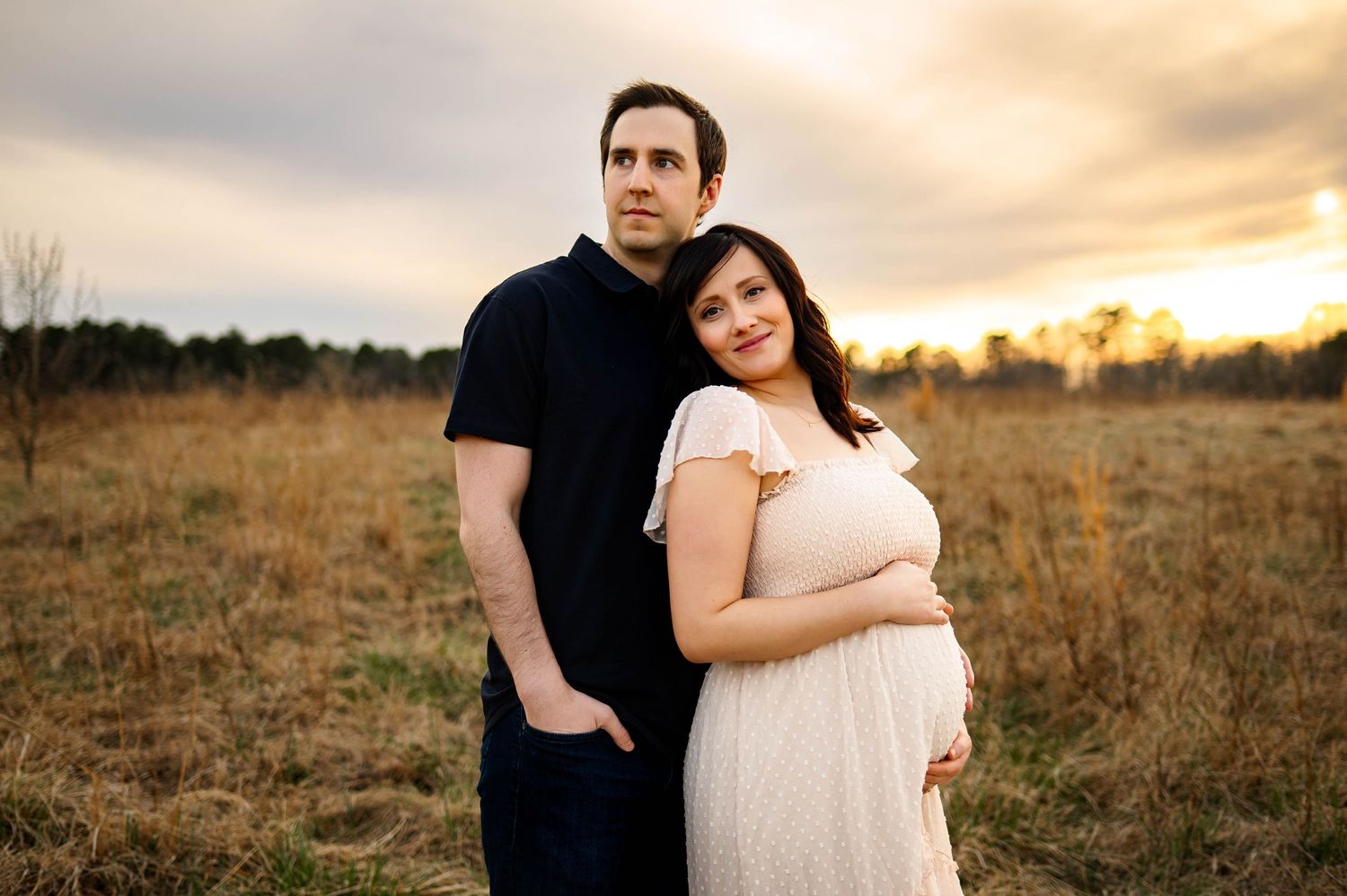 A couple embraces in a golden field at sunset, with mother wearing a flowing white dress with best wake forest maternity photographer
