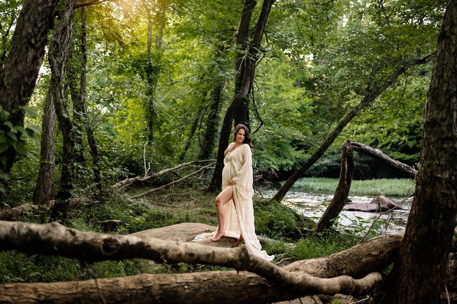 Mother in a flowing dress stands in a serene forest stream surrounded by lush green trees with maternity photographer chapel hill nc