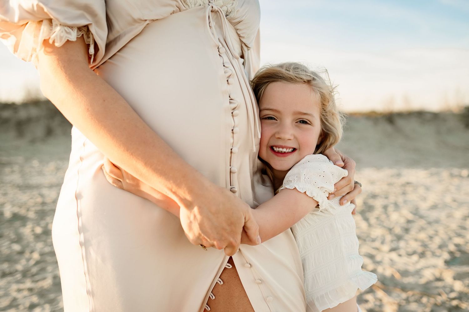 Young daughter hugs her mother's growing belly and smiles at the camera of a Raleigh maternity photographer