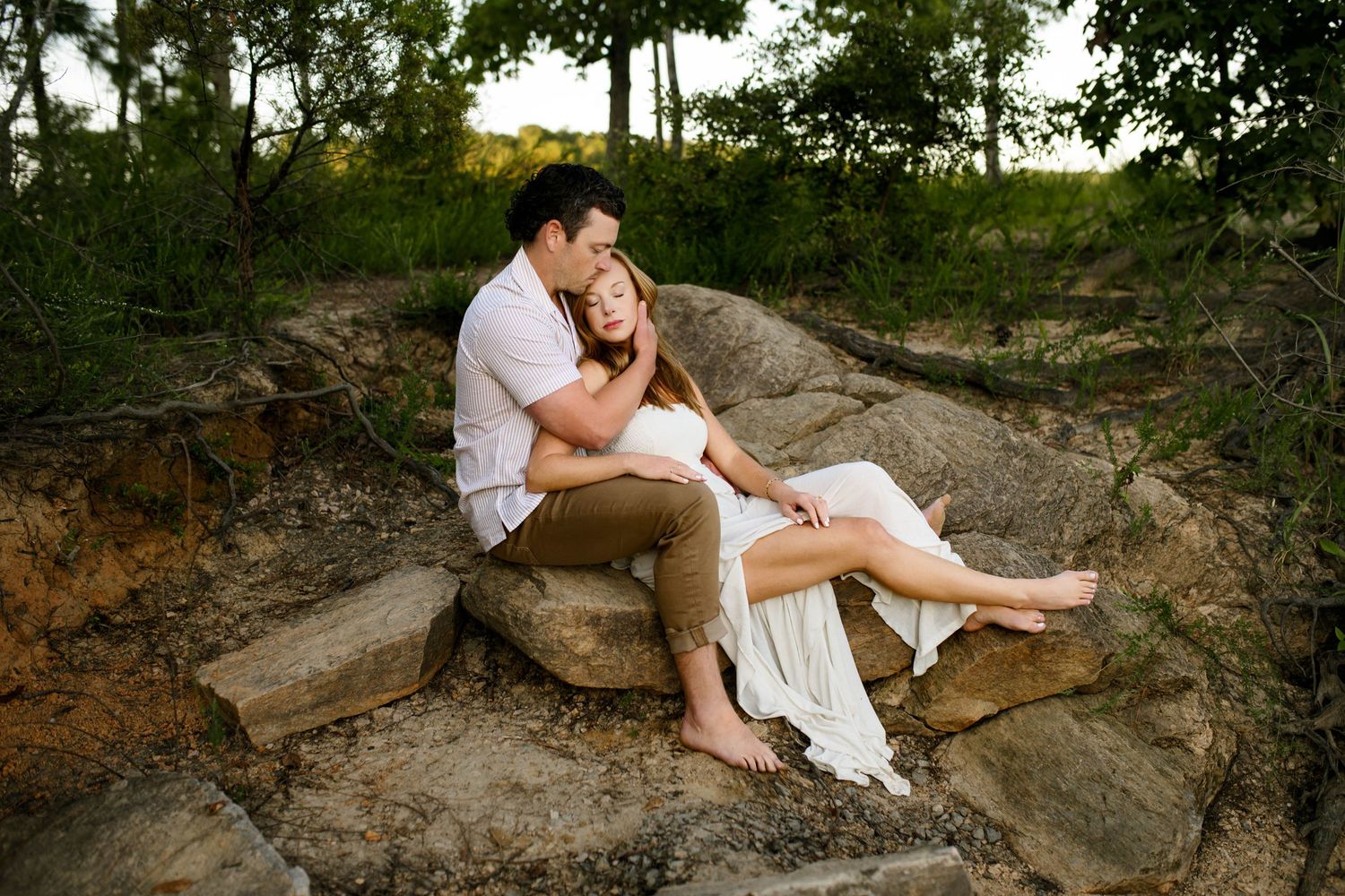 A couple in white outfits sits intimately together on large rocks in a natural outdoor setting during family photography session in Raleigh