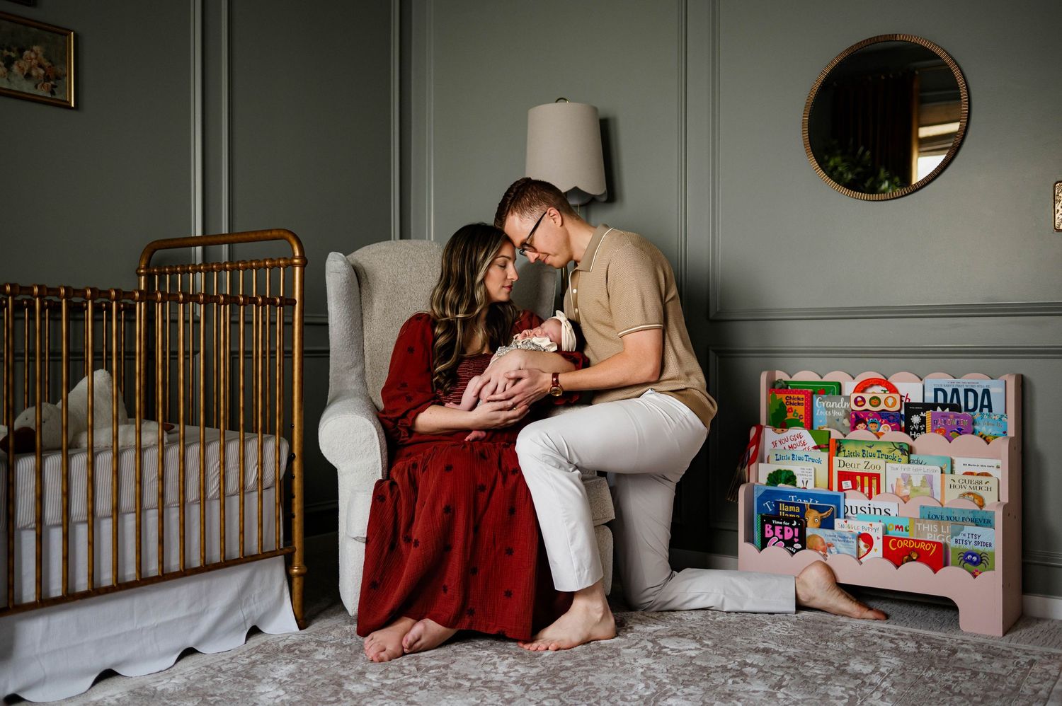 Parents sit together on a white chair next to a wooden crib and children's books in a cozy nursery with durham newborn photographer
