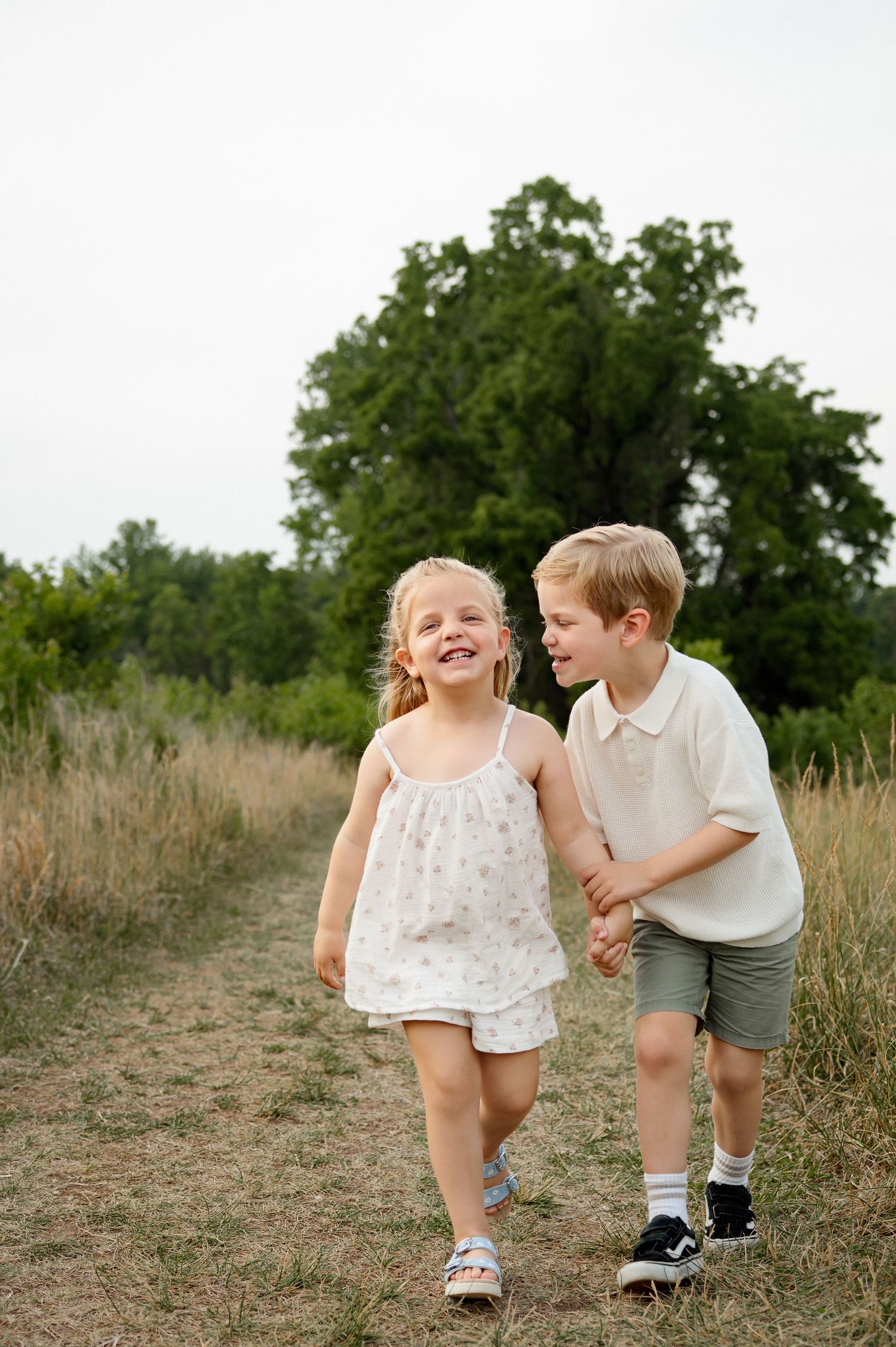 Brother and sister holding hands and walking along a path through tall grass during a Raleigh family photography session