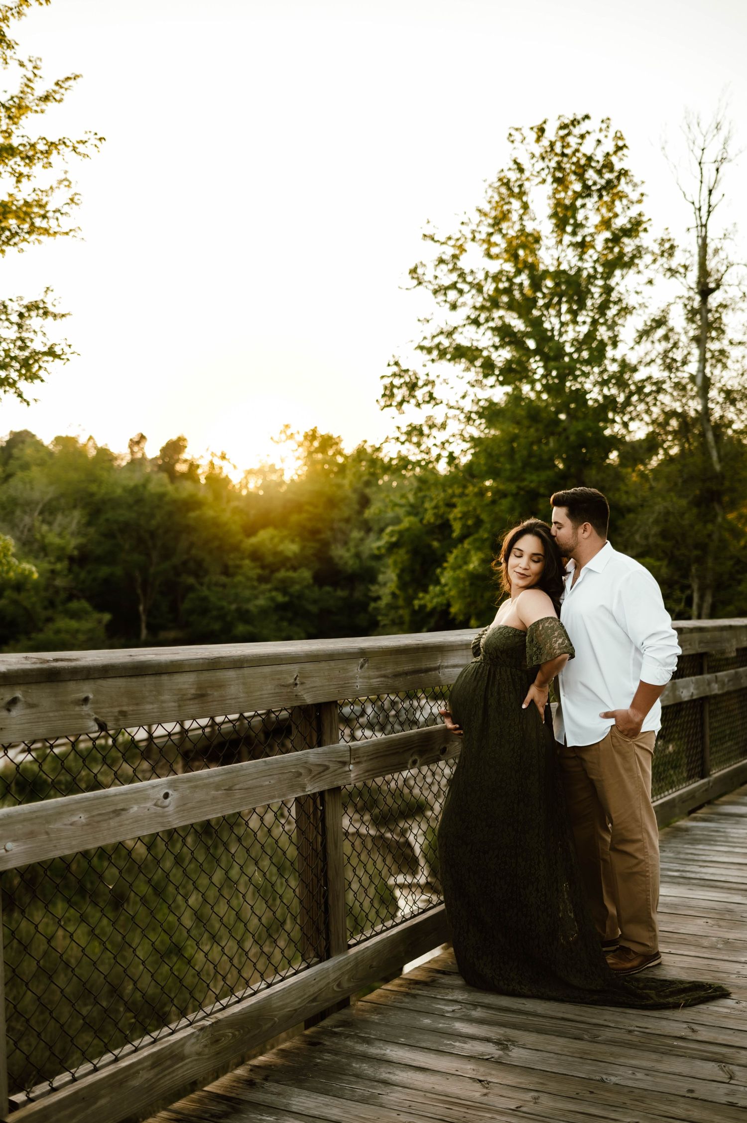 A couple shares a romantic moment on a wooden bridge at sunset as light filters through trees with raleigh maternity photographer