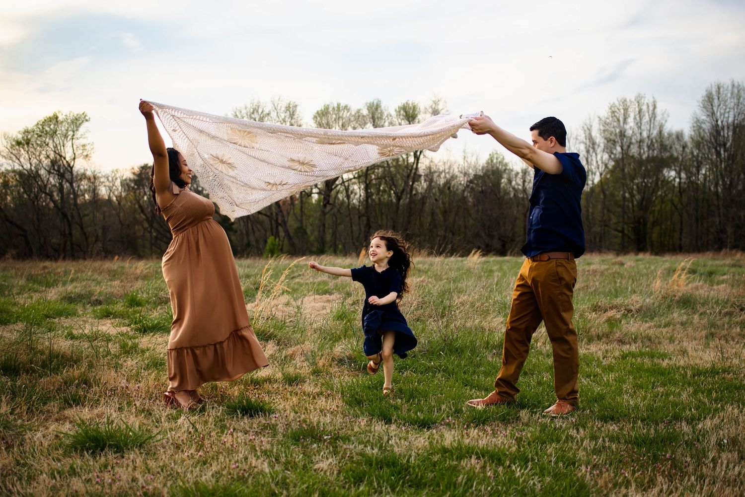 Family plays outdoors in a grassy field, holding up a white blankey at sunset with wake forest maternity photographer