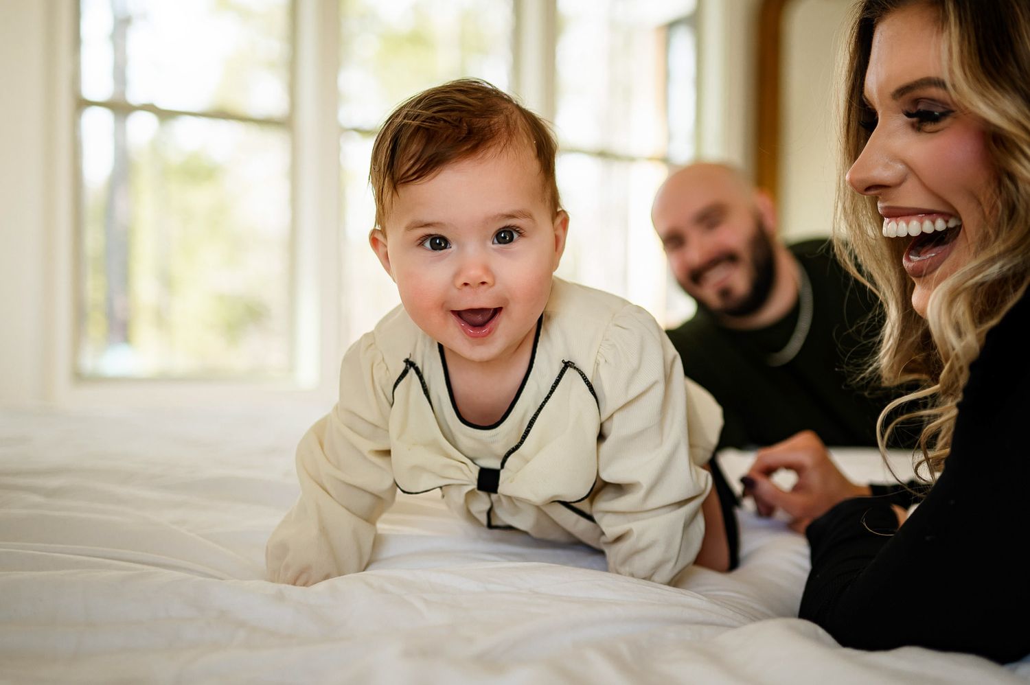 Baby crawling on a bed while parents smile in the background during an in-home family photography session in Raleigh