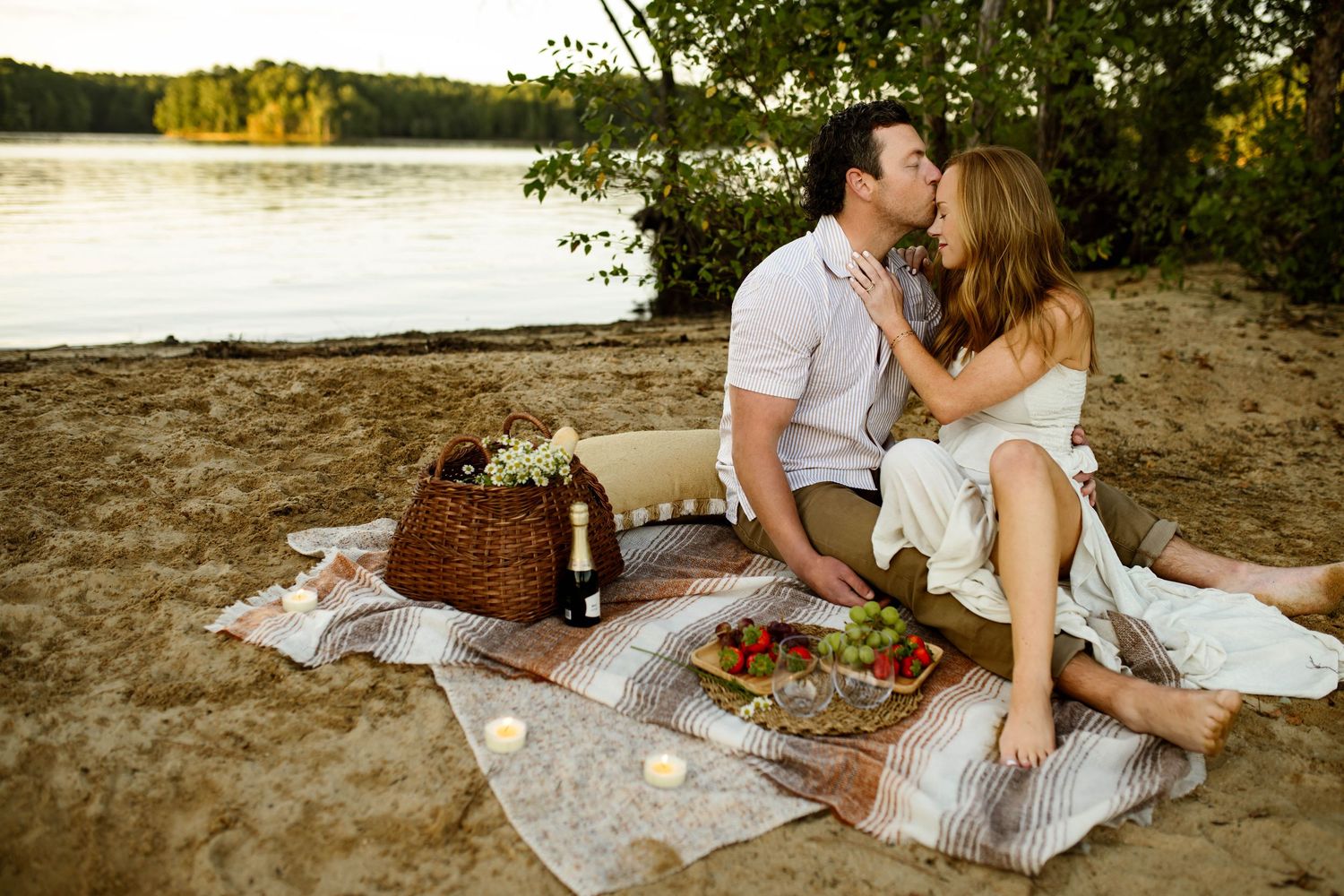 Romantic picnic scene on a sandy beach at sunset with wine, basket and blanket spread near tranquil lake waters with family photographer