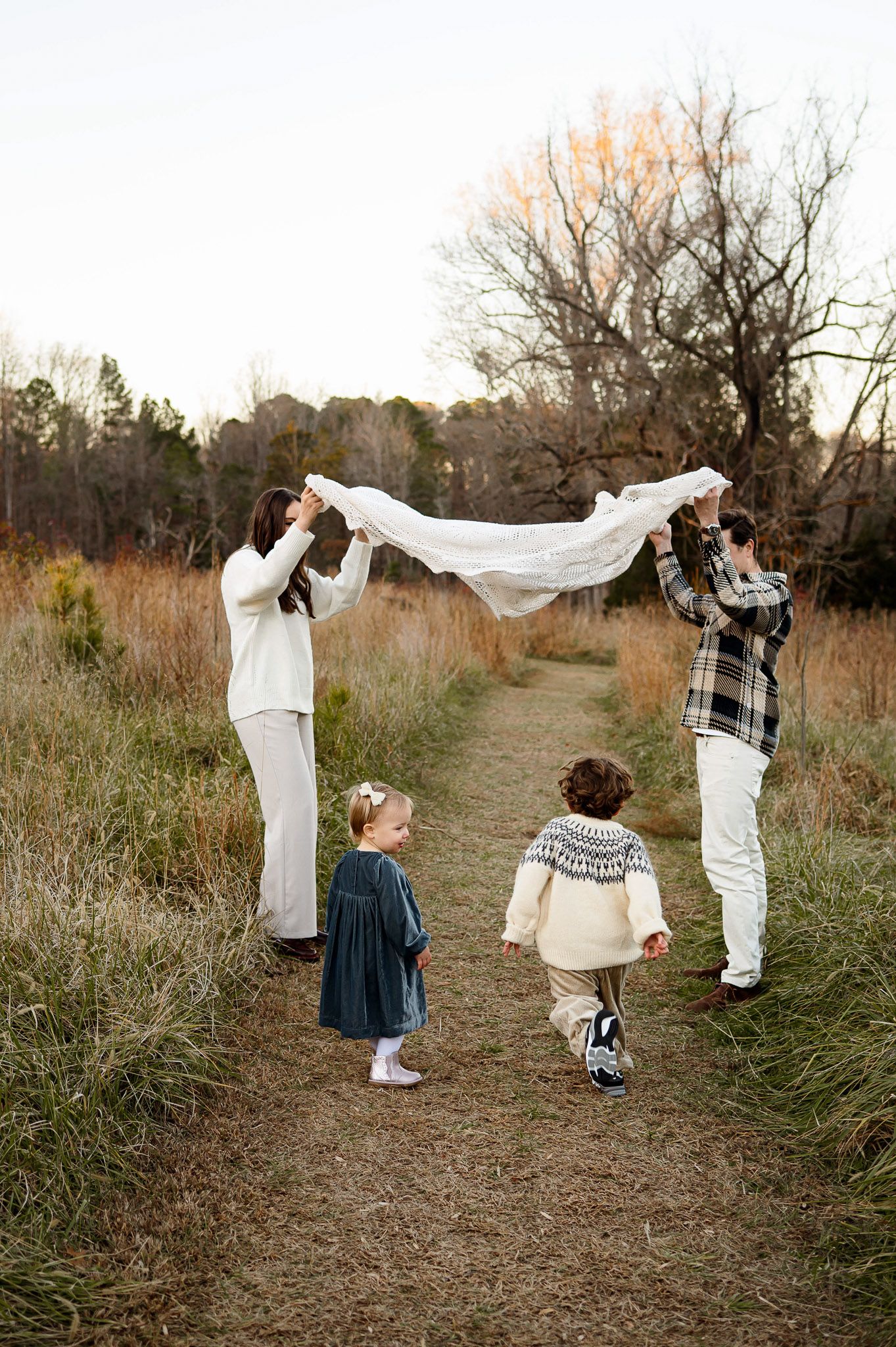 Parents holding a blanket while their children walk beneath it during a playful outdoor family photography session in Raleigh