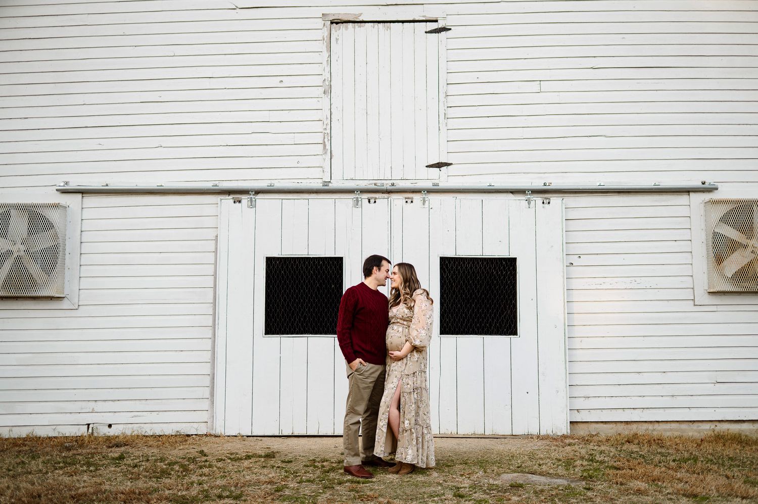 Expecting parents standing together in front of a white barn during an outdoor maternity photography session near Raleigh NC