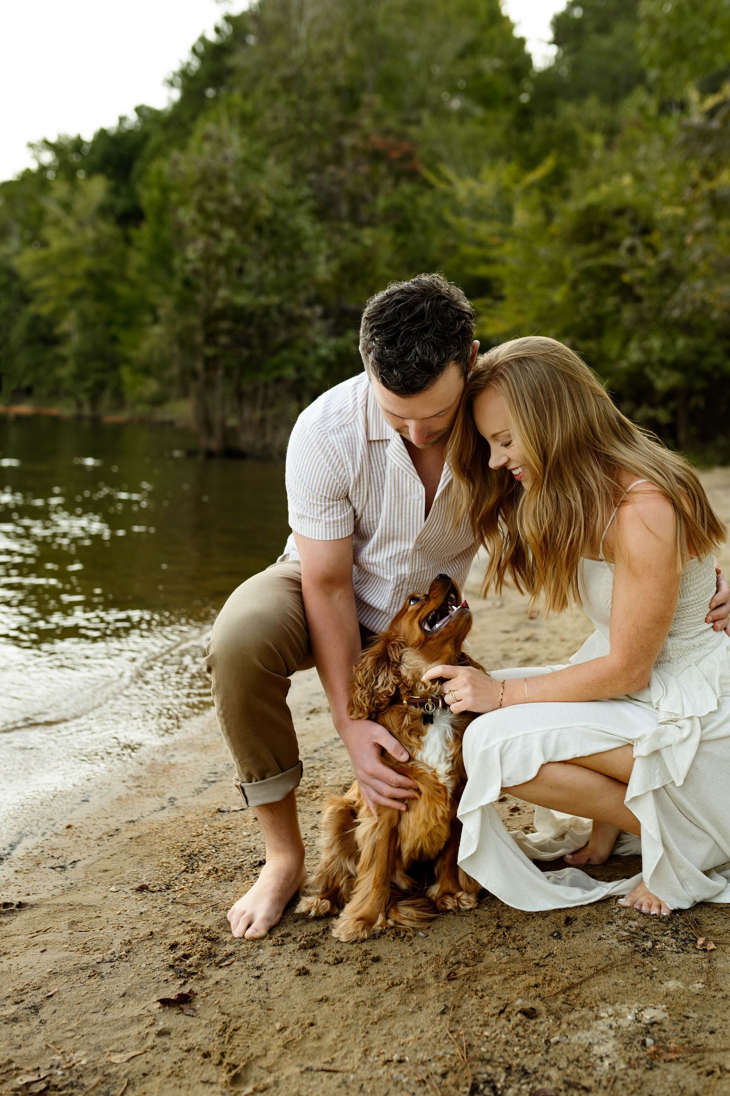 A couple in white clothing sits on a beach with their small brown dog enjoying a tender moment by the water with Raleigh family photographer