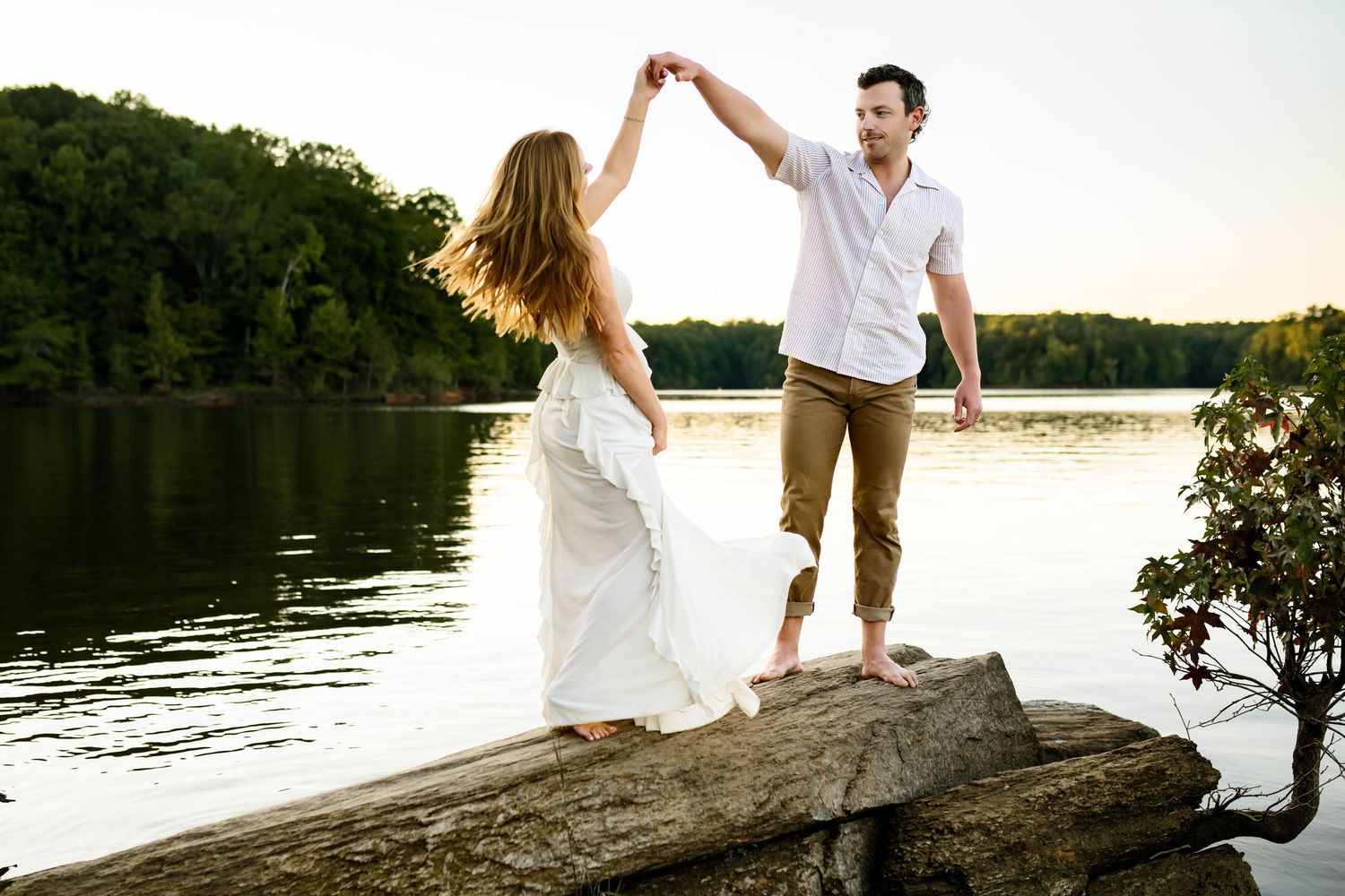 A couple in white clothing stands together on a wooden dock by a serene lake at sunset with family photographer Emma Branch Photography