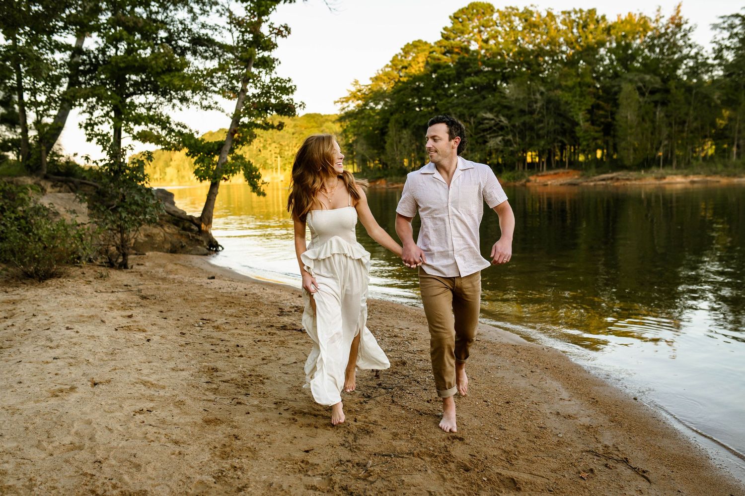 A couple in white and brown attire walks along a scenic waterfront path during golden hour sunset with Raleigh newborn photographer