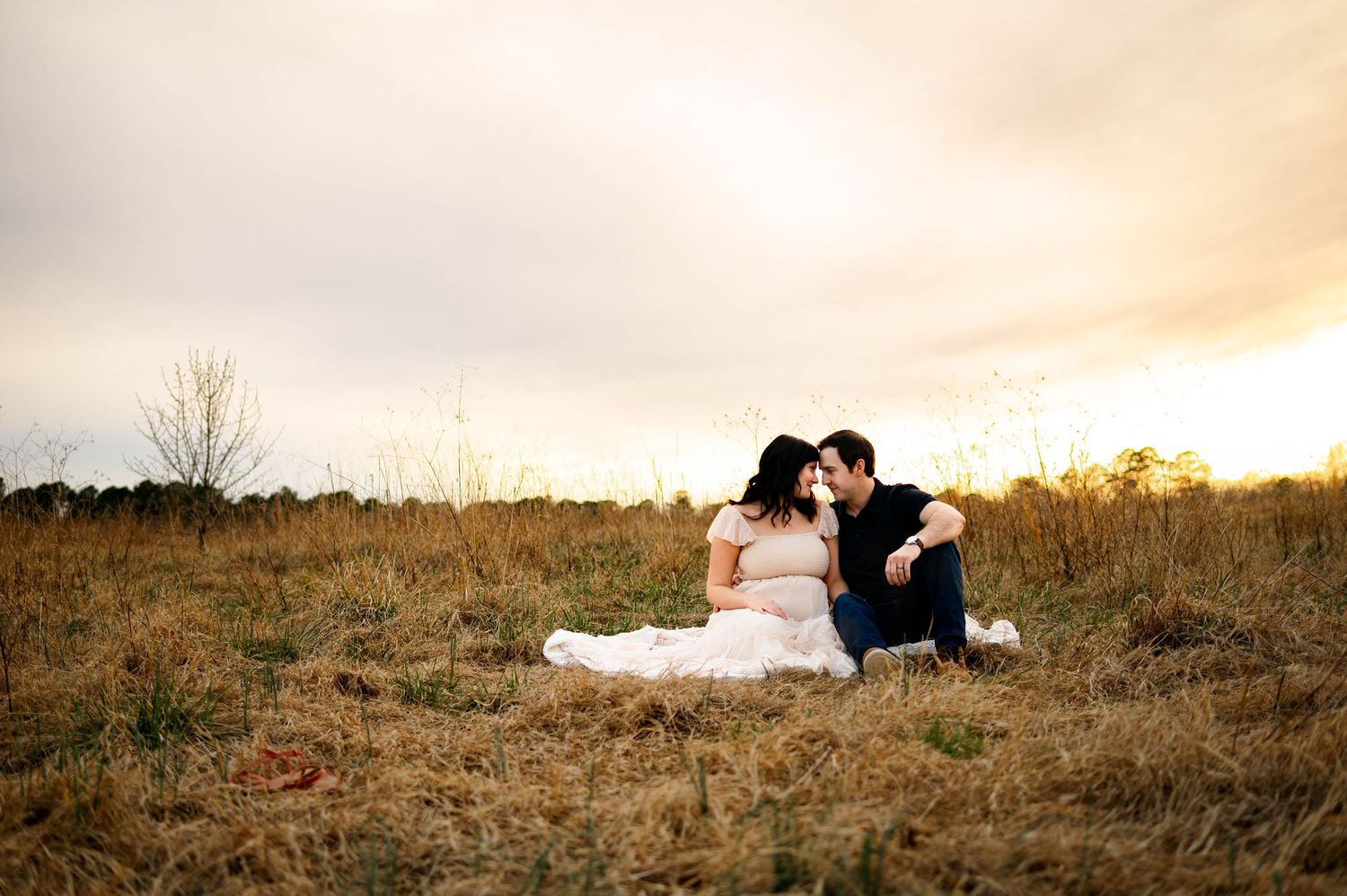 A silhouetted couple sits together in a grassy field during golden hour sunset with best maternity photographer Raleigh