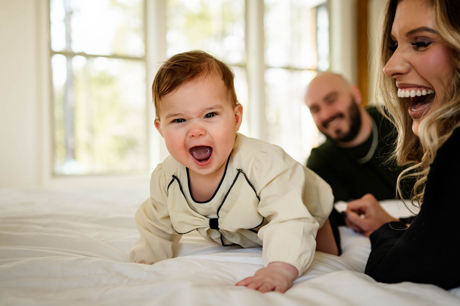 A joyful baby in cream outfit crawls on white bedding while parents smile in the background for an in home photography shoot raleigh nc