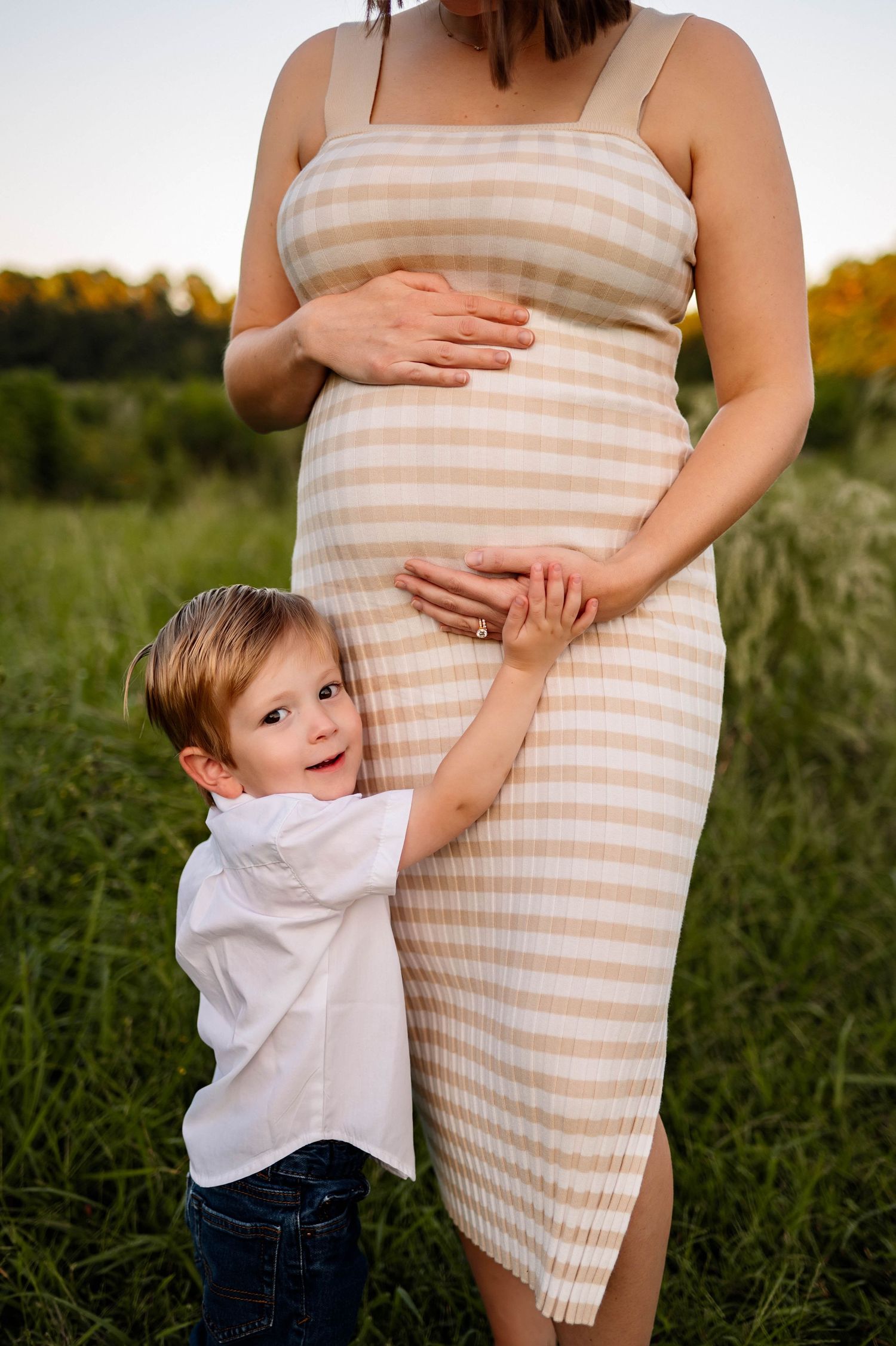 A child in a white shirt hugs his mother's pregnancy bump while standing in a grassy field at sunset for maternity photography in raleigh nc