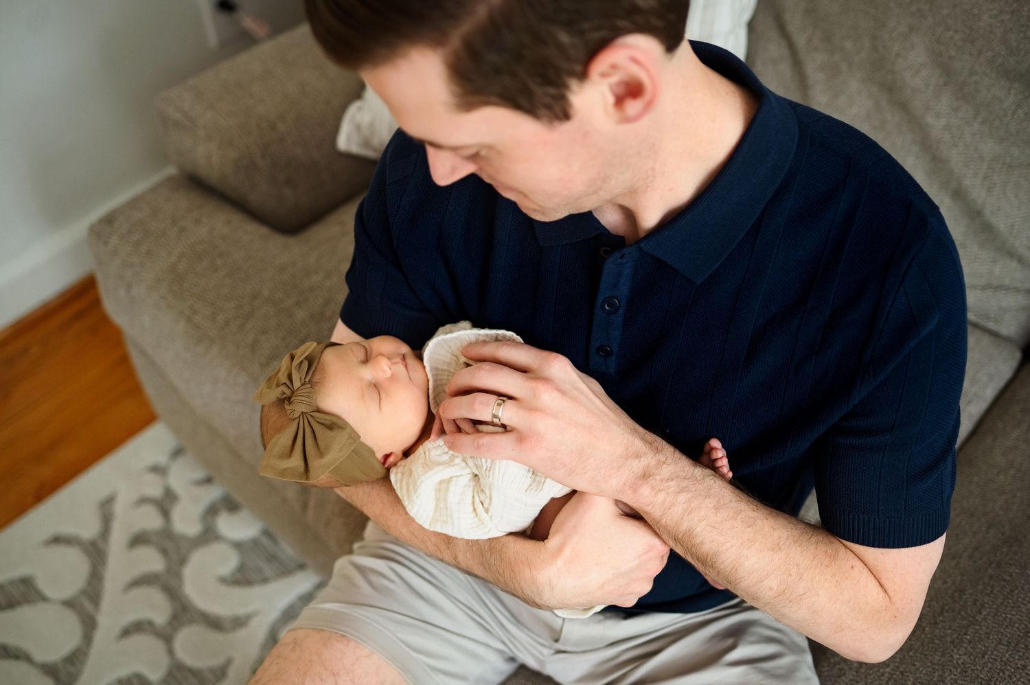 Father admires newborn daughter while holding on couch in living room during a photoshoot with a Raleigh family photographer