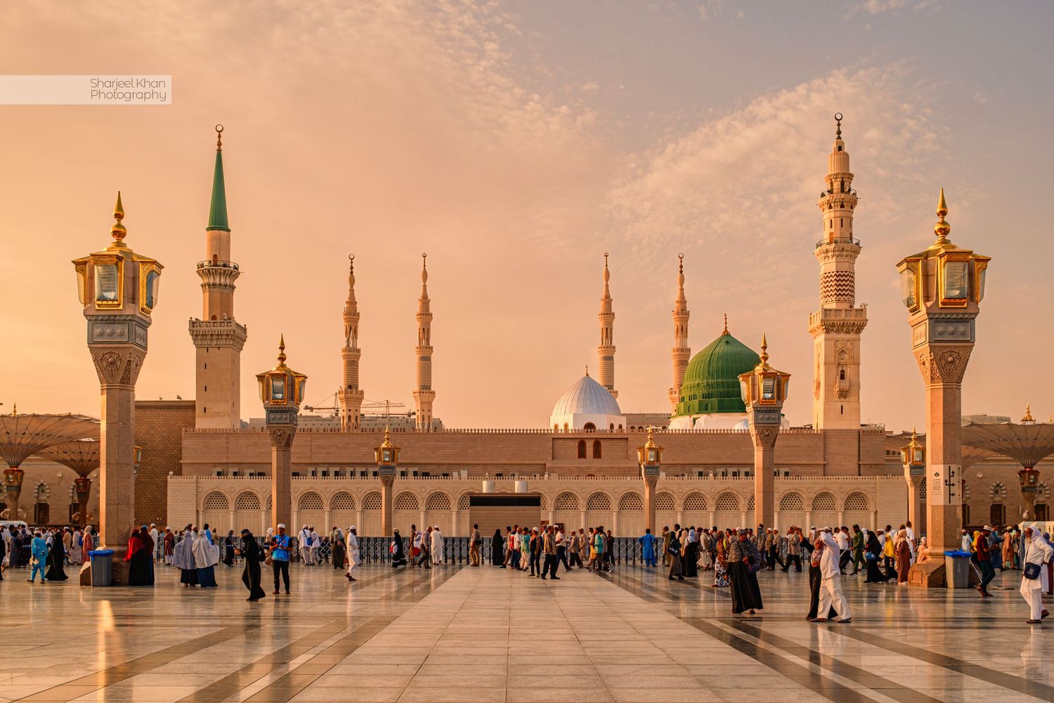 A panoramic view of the Prophet's mosque's courtyard in Madinah, KSA, with ornate minarets and worshippers during the golden sunset hour.