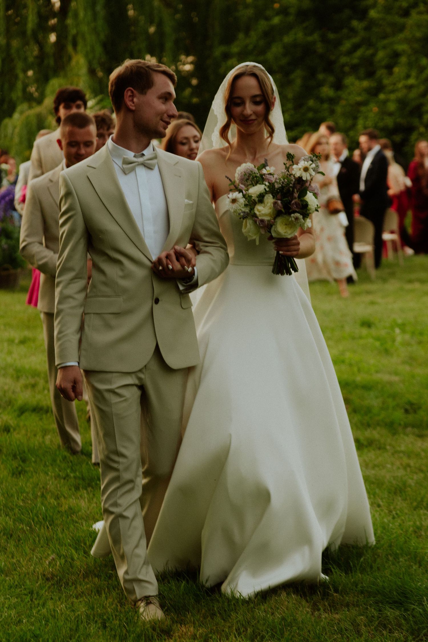 Wedding couple walks across lush green lawn with bridal party, groom in tan suit and bride in flowing white gown.