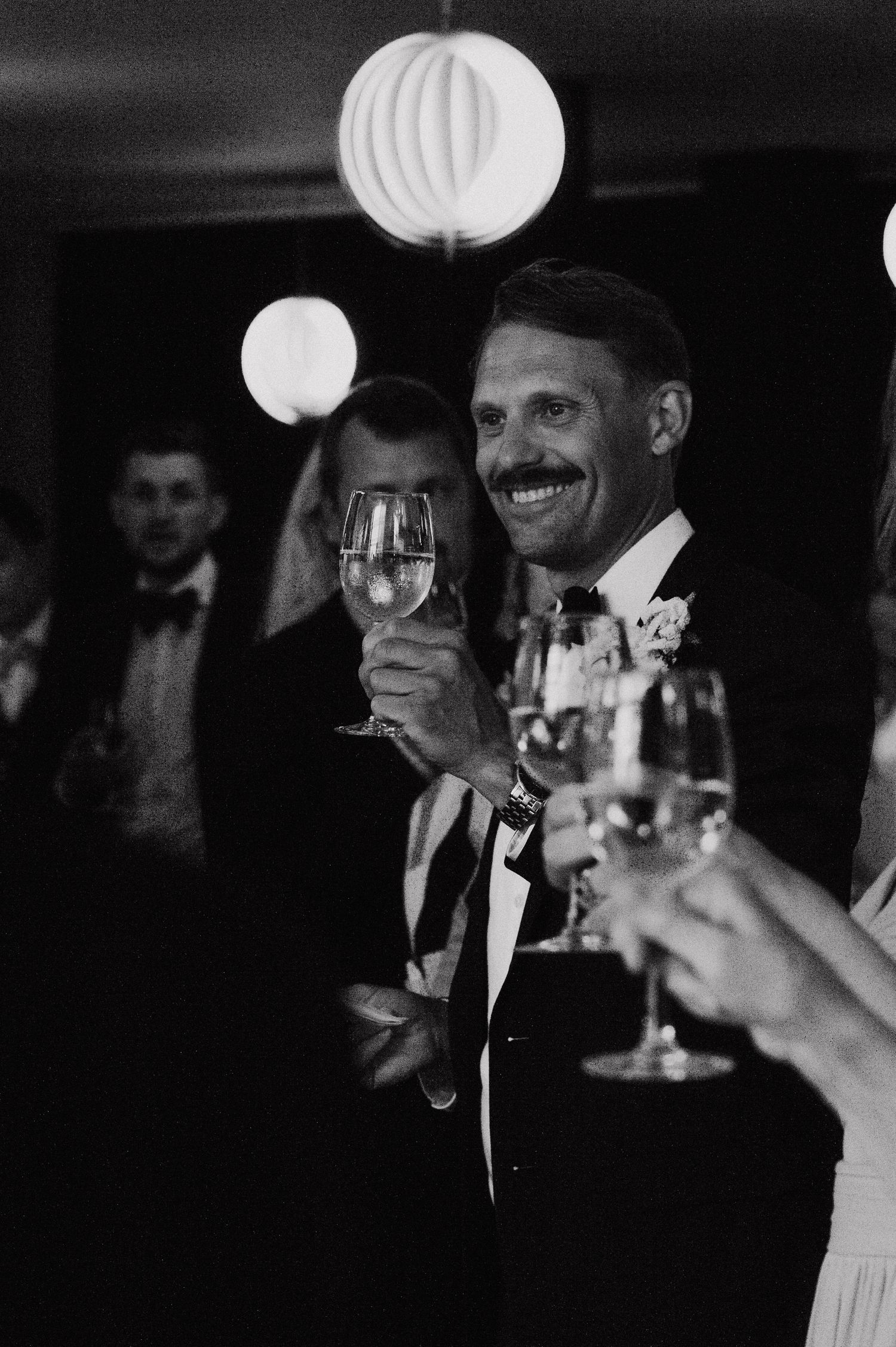 Black and white photo of wedding guests raising wine glasses in a toast during reception.