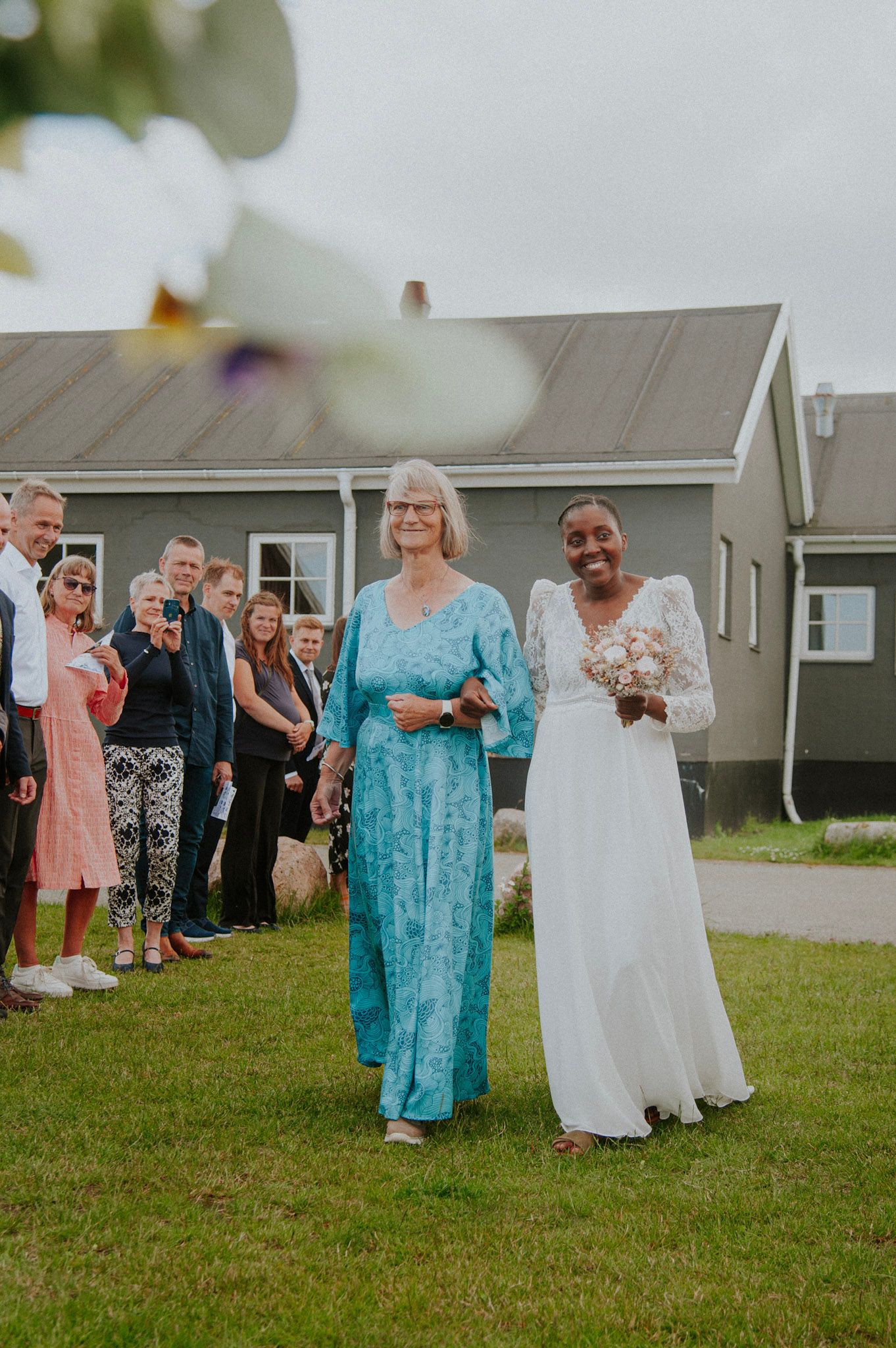 Wedding guests in colorful attire gather on a green lawn outside a coastal venue.