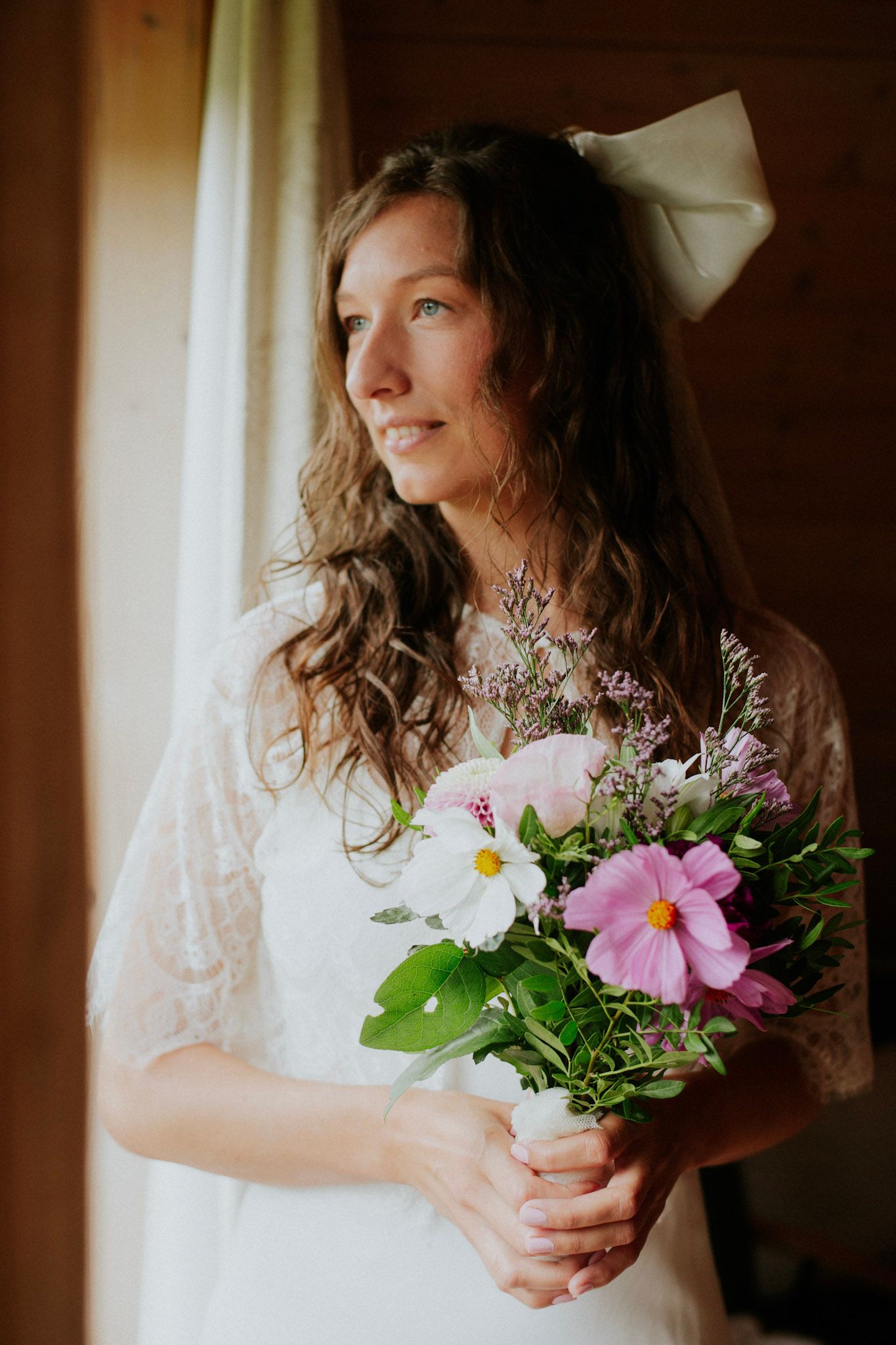 Bride in lace dress holding delicate bouquet of pink and white flowers by window light.