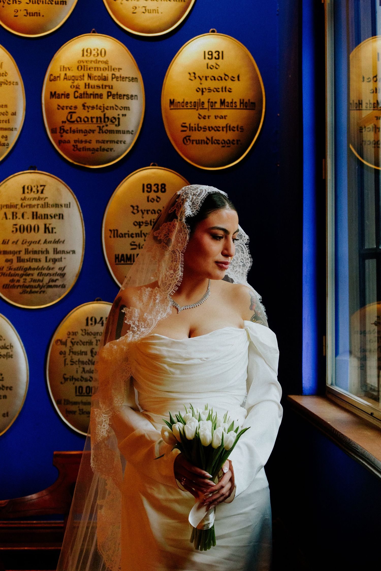 Bride in off-shoulder dress poses with white tulip bouquet against blue wall with golden plaques.