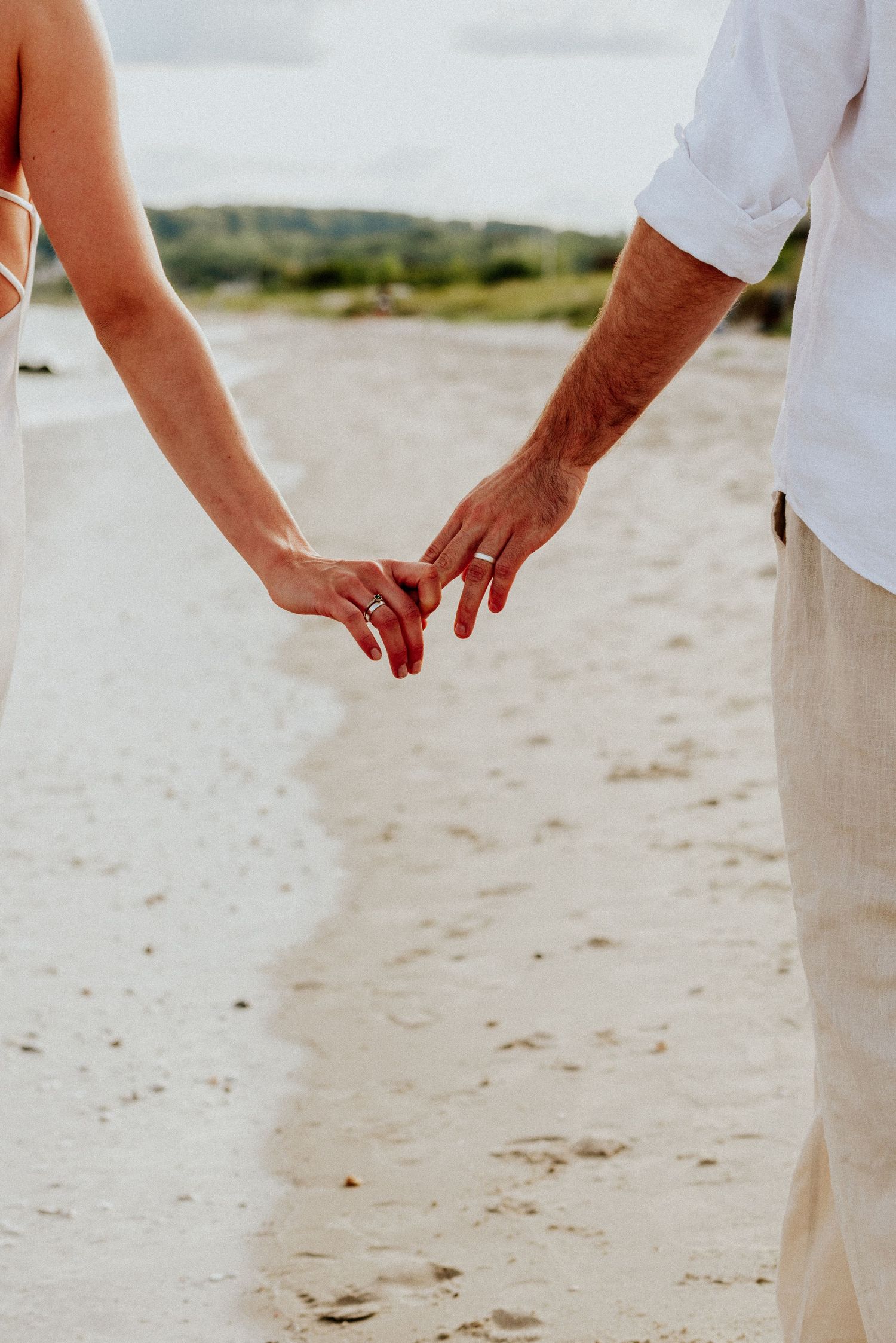 A couple holds hands tenderly while walking on a sandy beach, wearing white wedding attire.