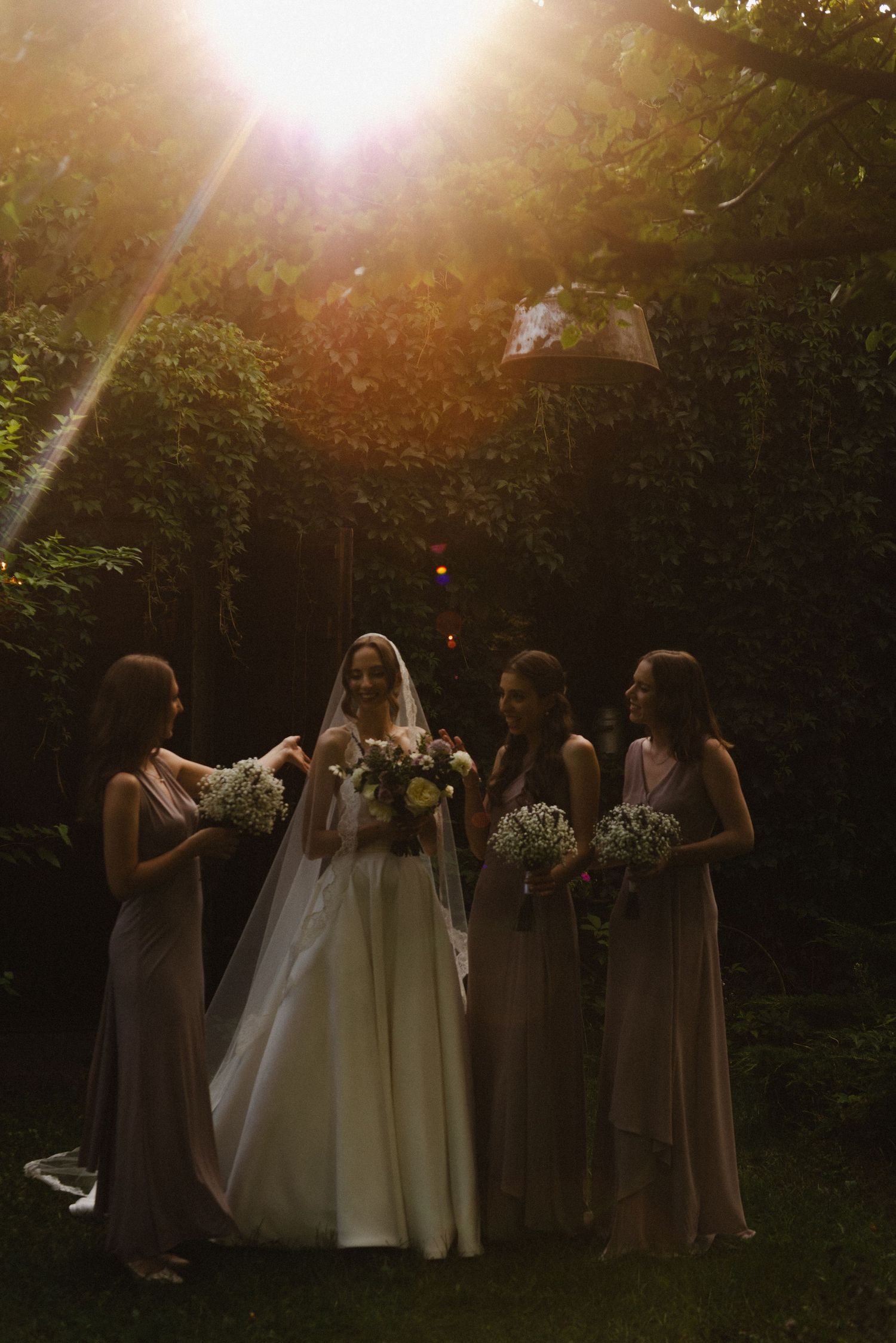 Wedding party members in mauve dresses gather around bride in white gown during sunset in garden setting.