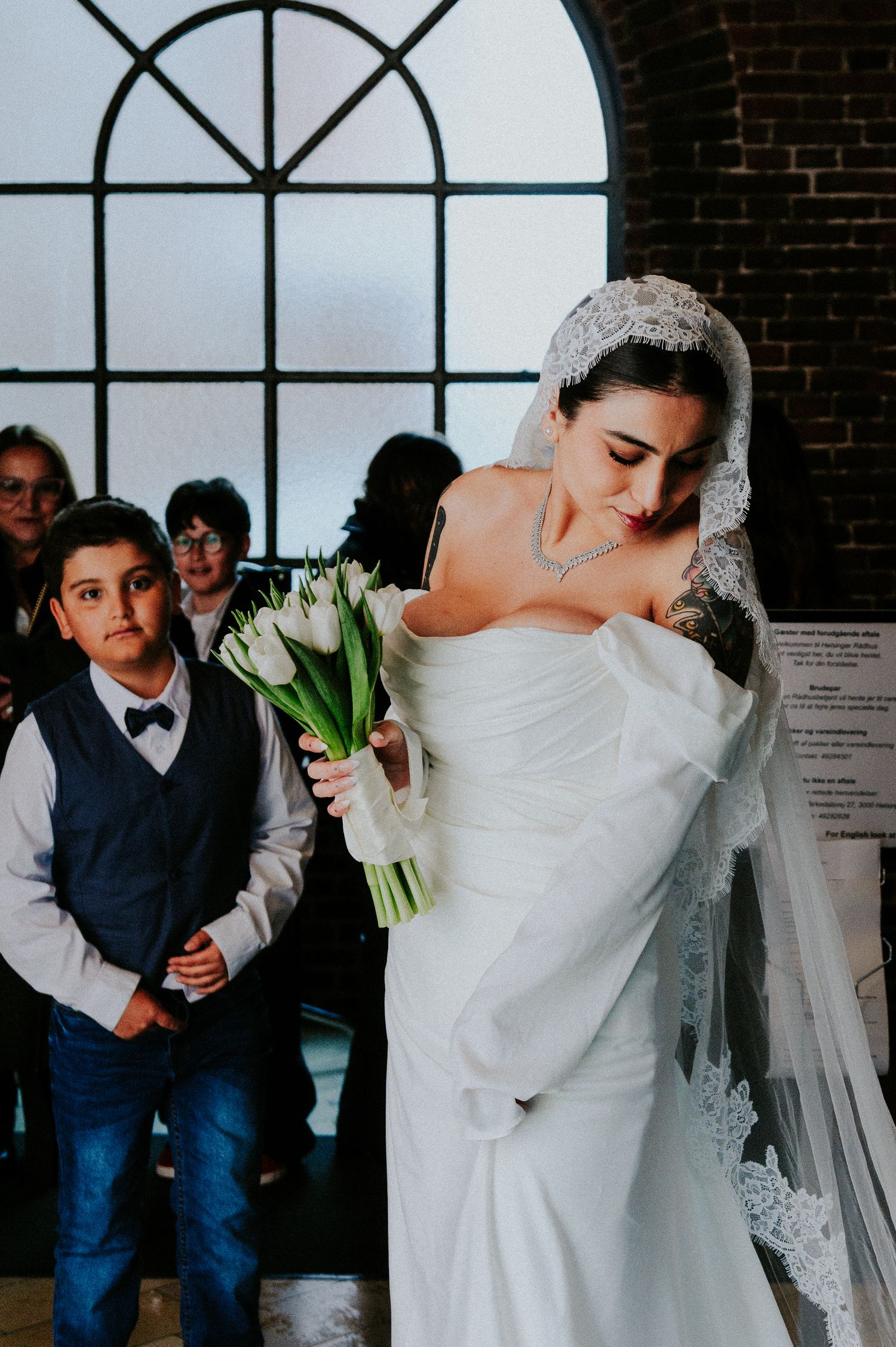 Elegant bride in an off-shoulder white gown holds white tulips while standing by a large arched window.
