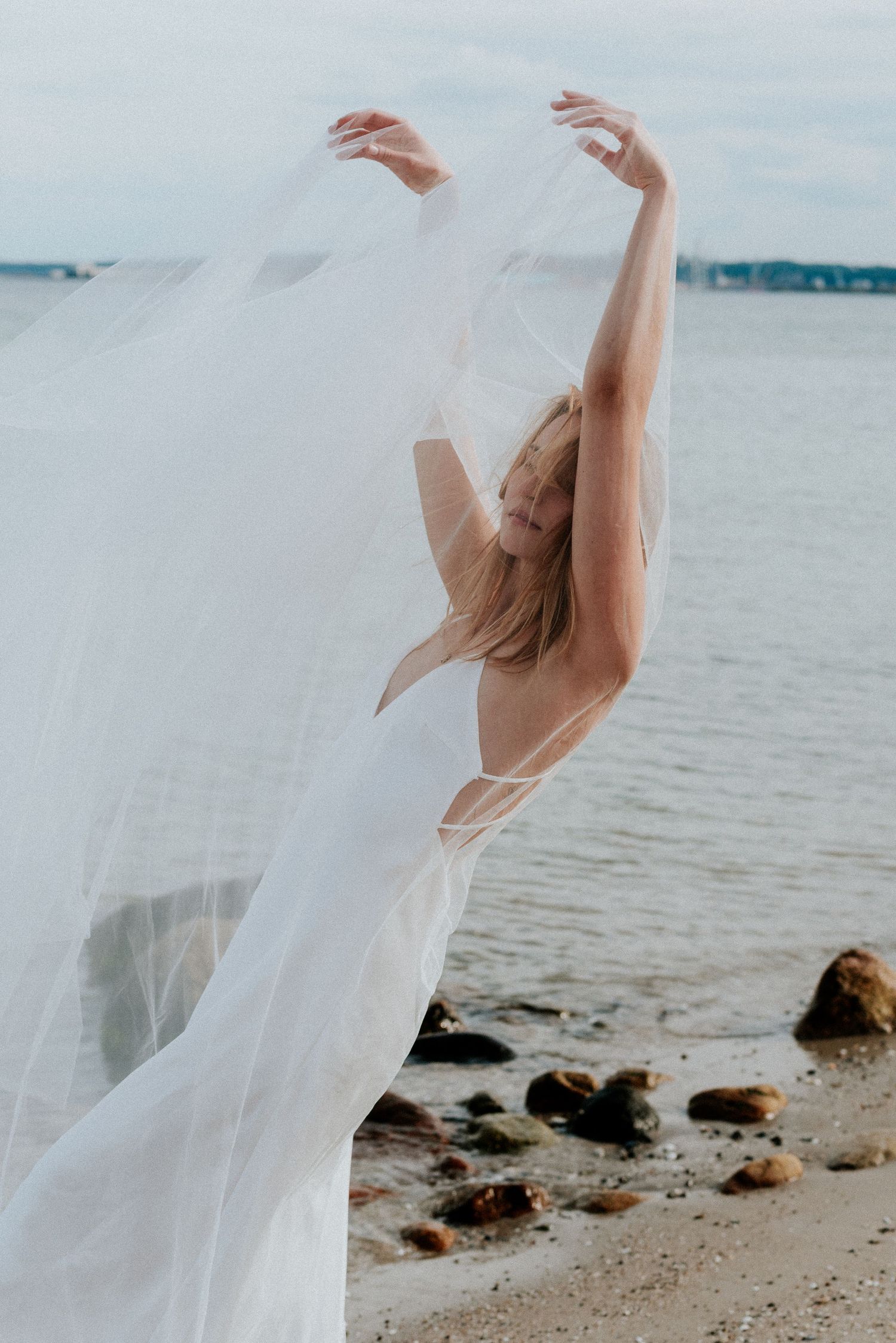 A graceful dancer in a flowing white dress poses elegantly on a rocky beach shore with water in the background.