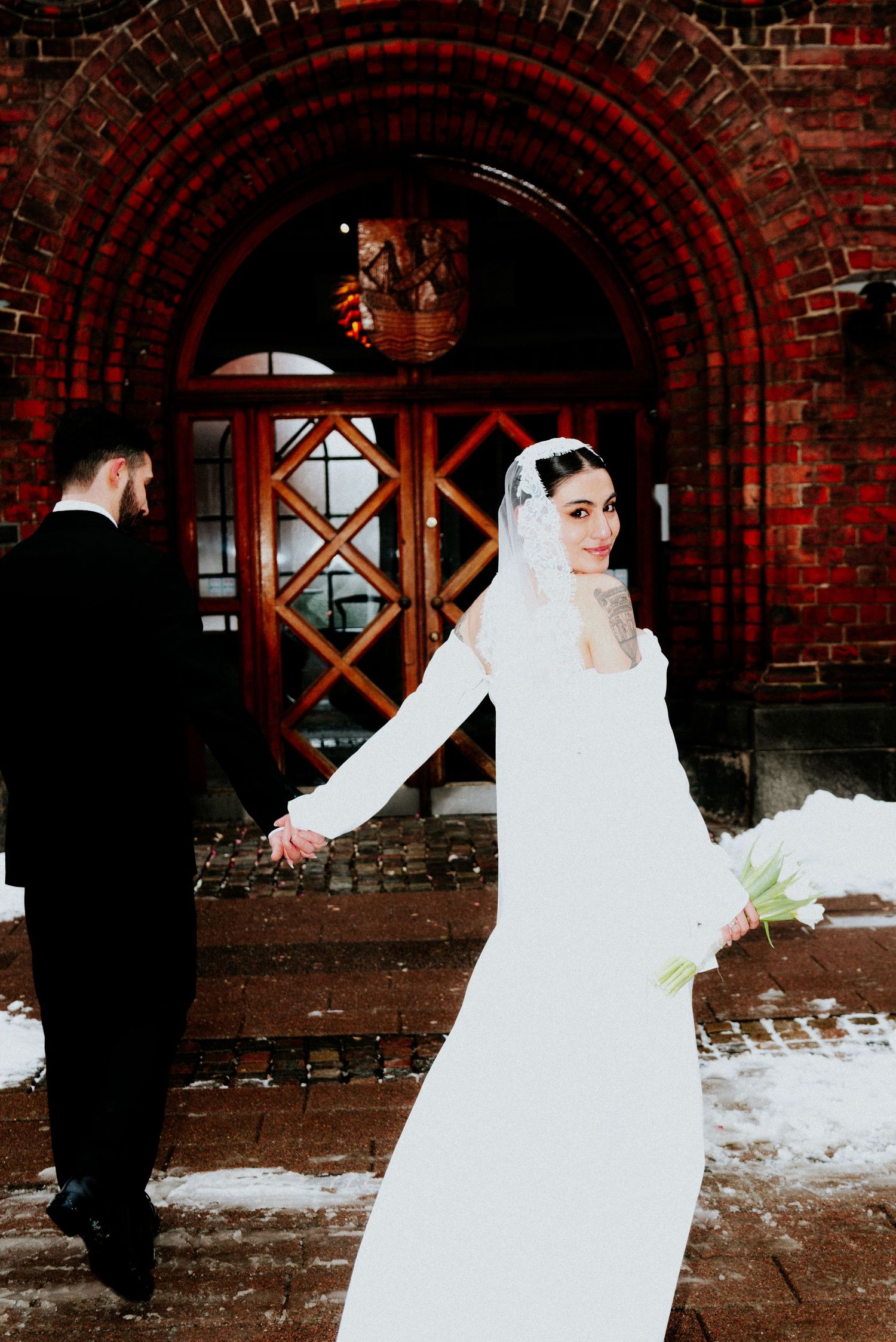 Winter wedding scene with couple in formal attire against red brick archway entrance.