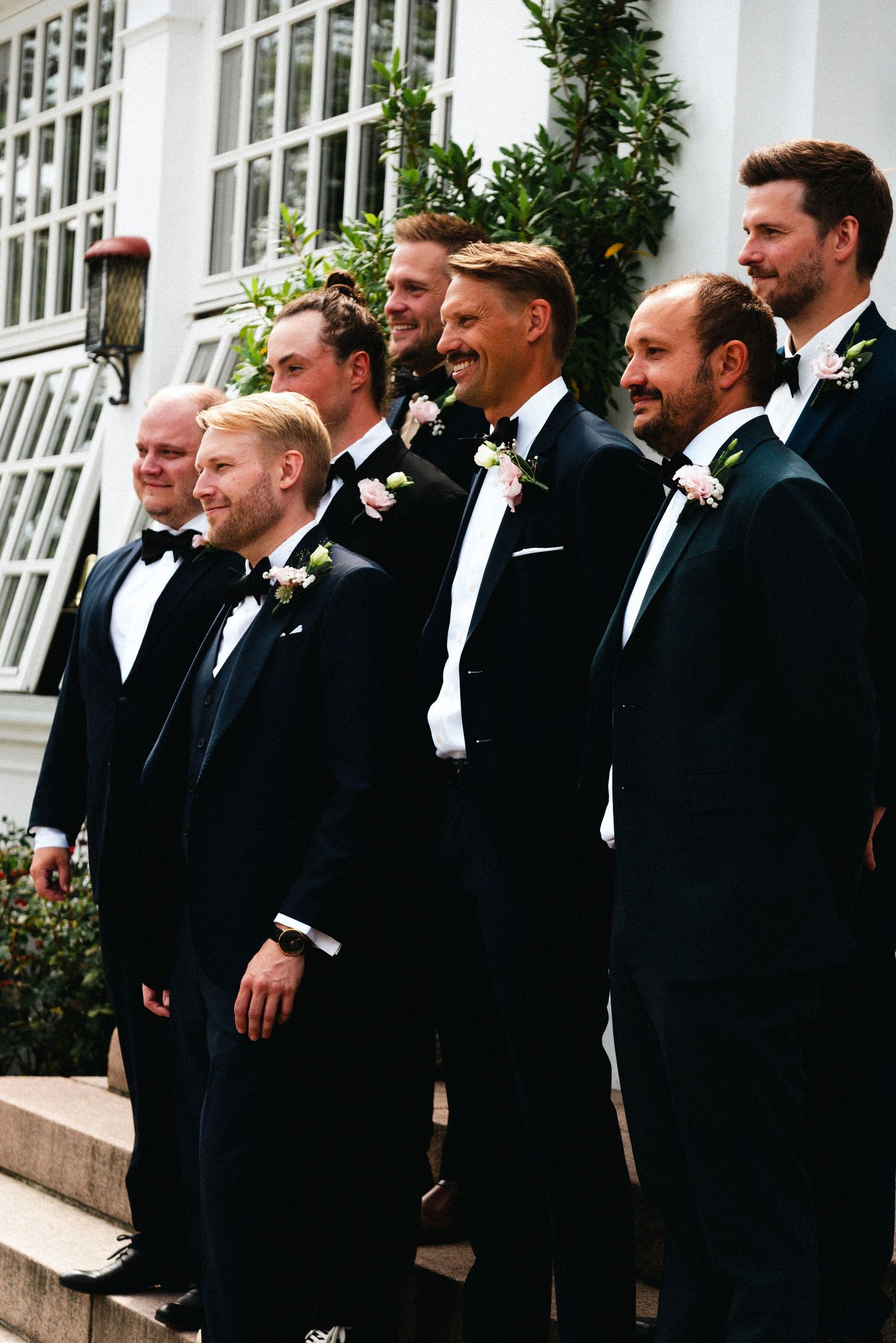 Groomsmen in black tuxedos and bowties stand together smiling during a formal wedding photo session.