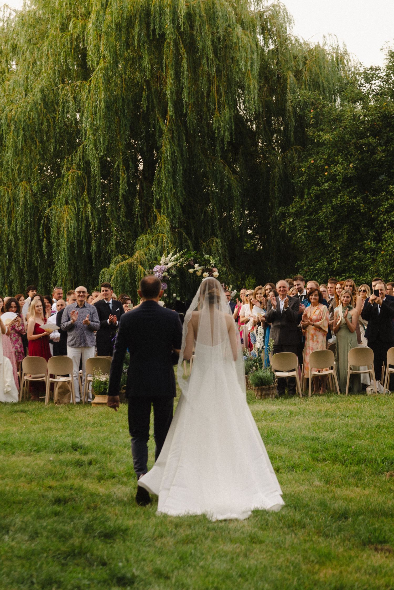 A wedding ceremony takes place under a weeping willow tree with guests gathered on the lawn.