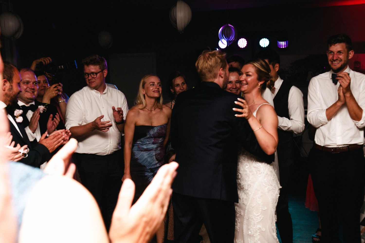 A group of people dancing and celebrating at a wedding reception in a dimly lit venue.