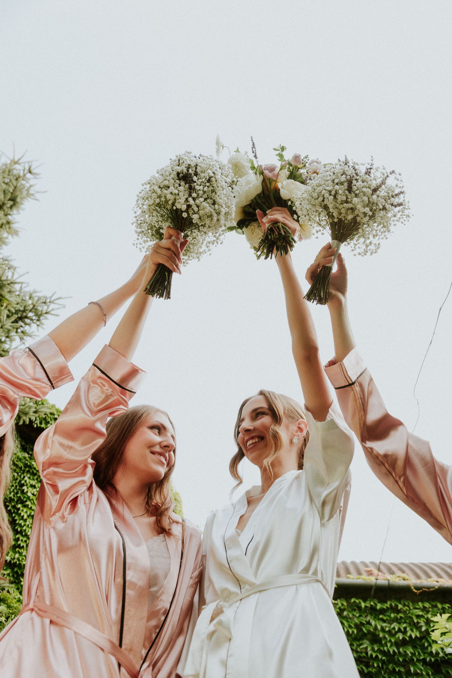 Bridesmaids in pink dresses and bride in white raise their bouquets joyfully during outdoor wedding celebration.