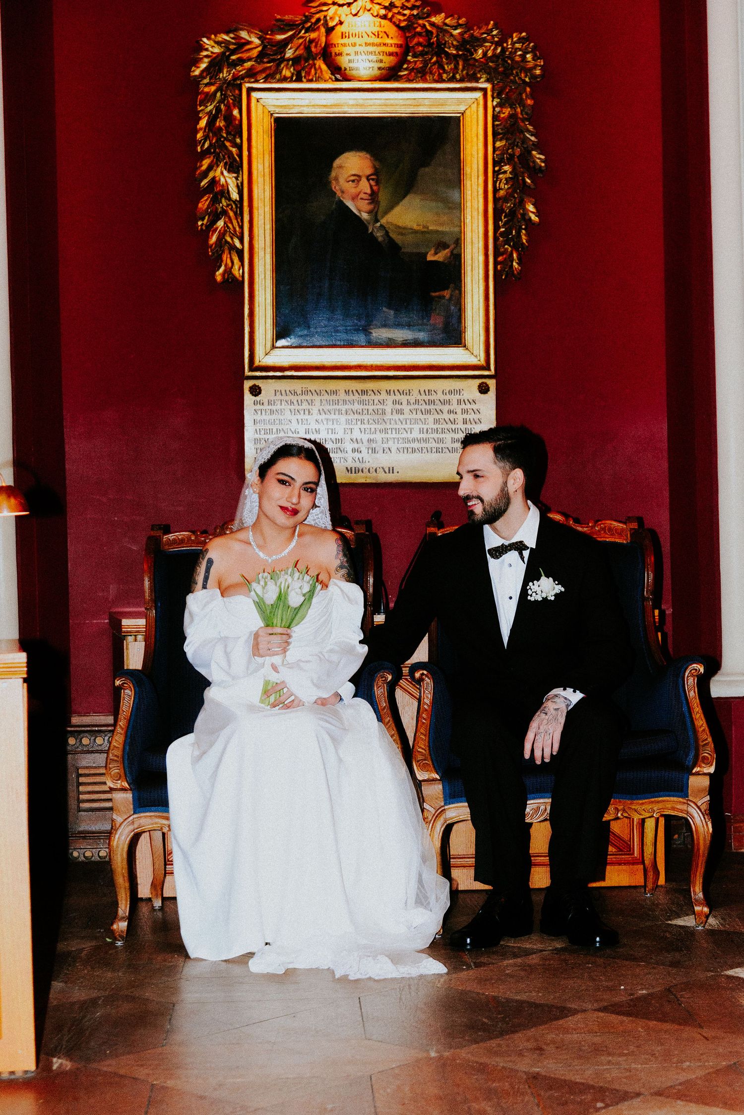 Wedding couple sits on elegant chairs in a grand room with red walls and ornate gold-framed portraits.