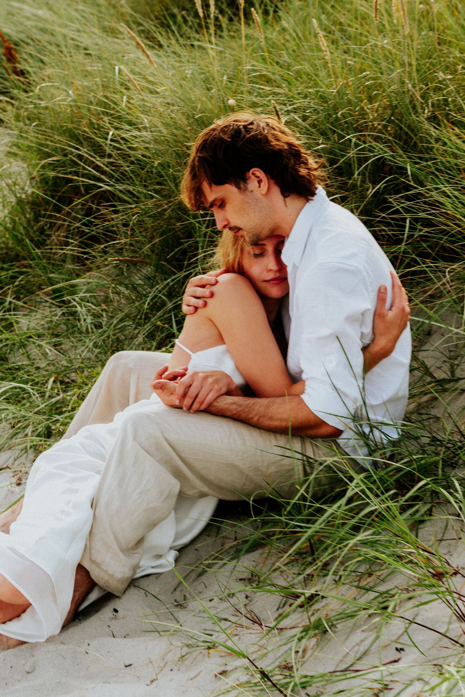 Romantic couple in white clothing embraces tenderly while lying together among beach grass dunes.