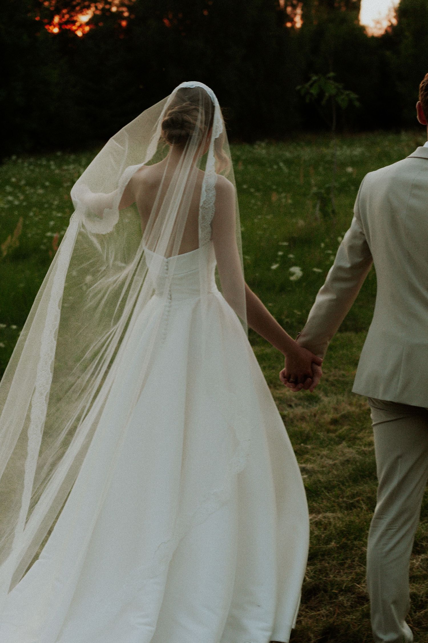 A romantic wedding scene with a bride in a flowing white dress and veil walking alongside someone in white outdoors.