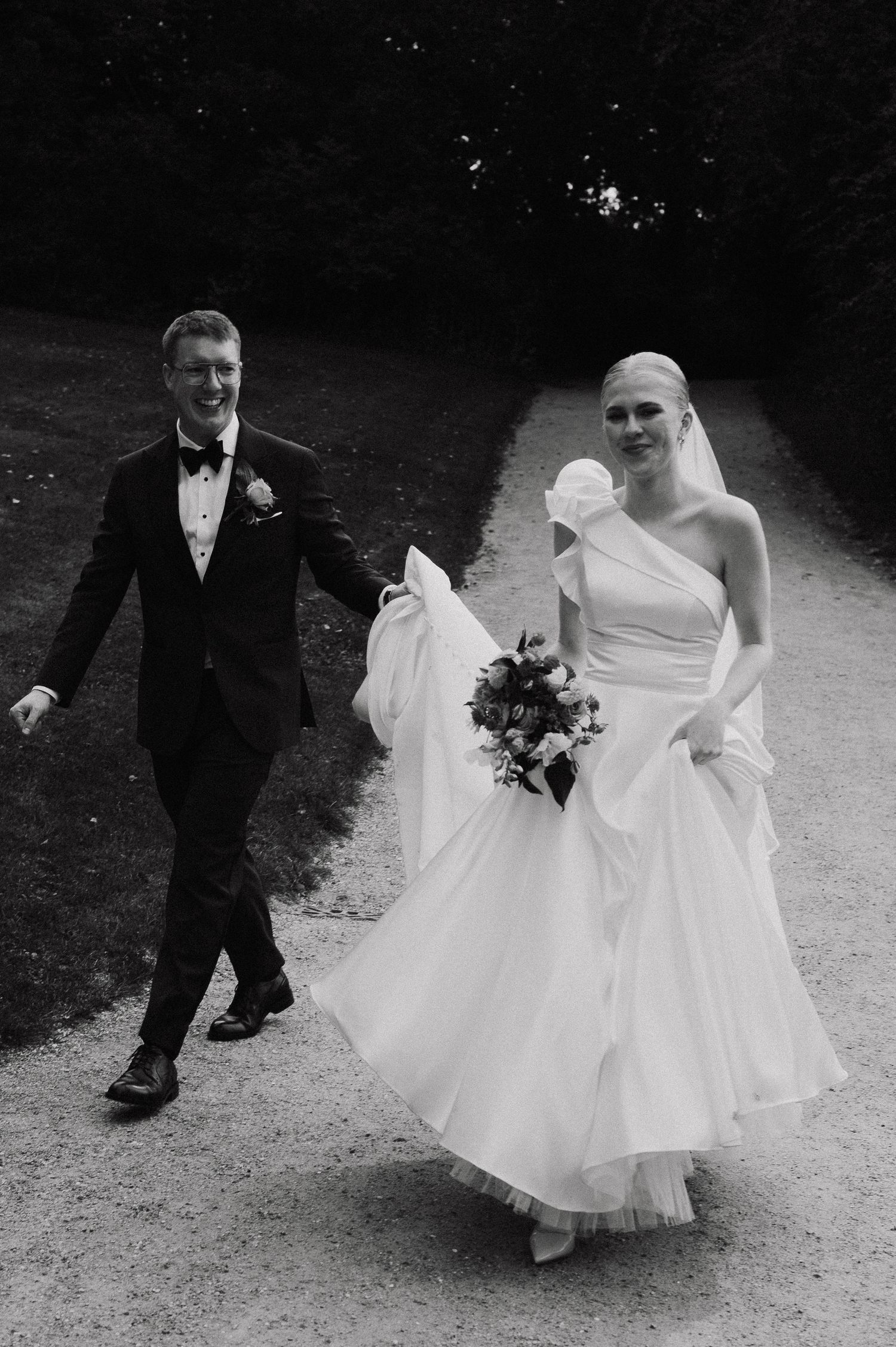 Black and white photo captures playful movement of bride's dress during outdoor wedding.