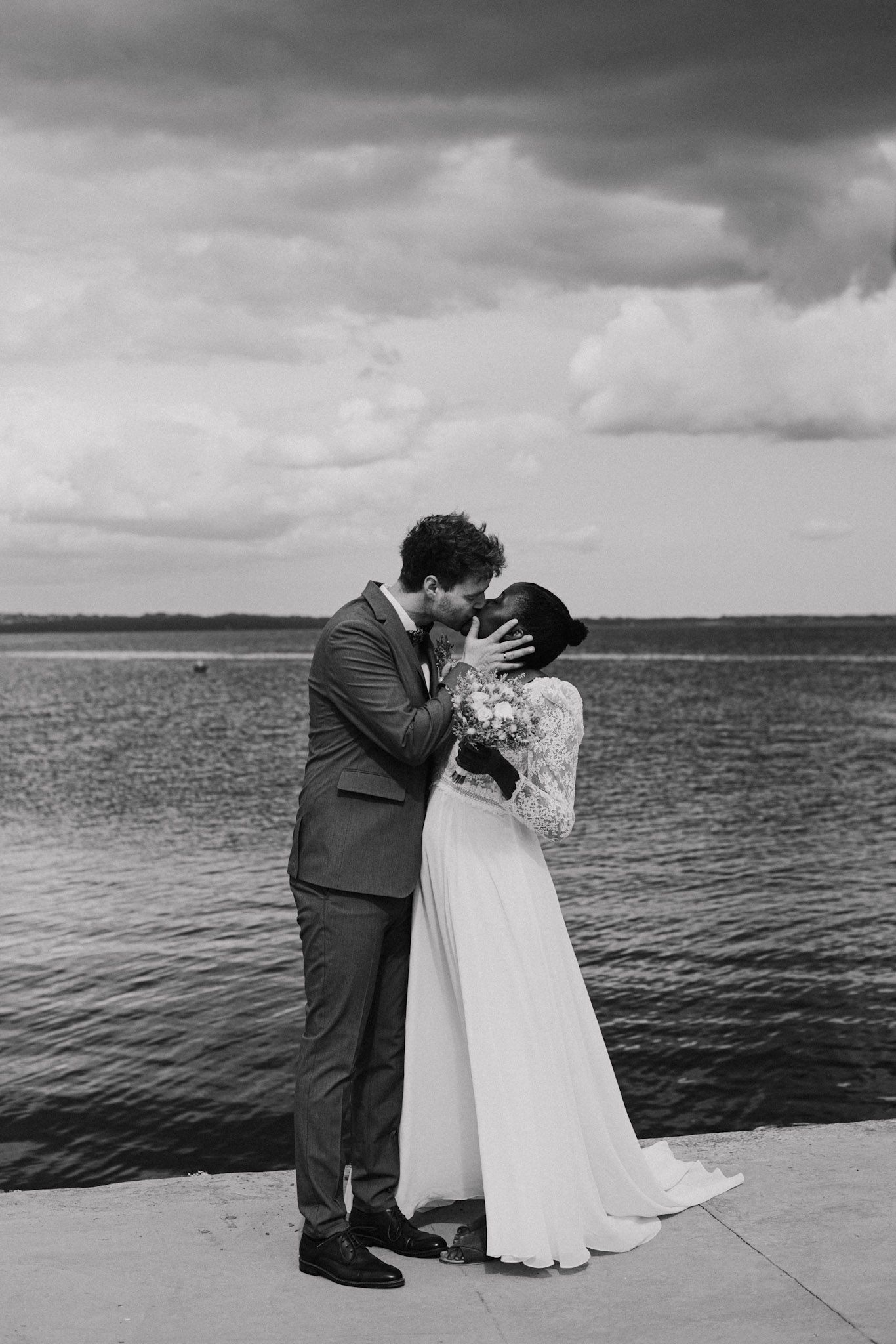 A romantic black and white wedding photo of a couple kissing by a lake with dramatic clouds overhead.