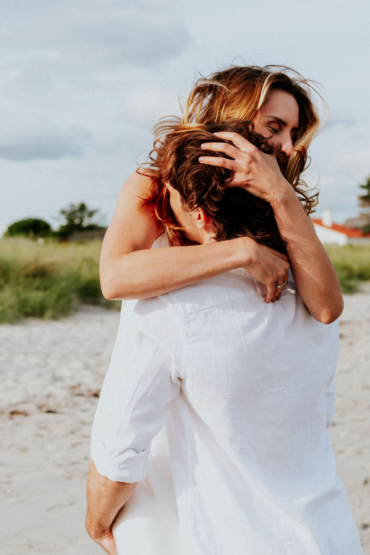 Two people wearing white embrace in a tender moment on a sandy beach with soft lighting.