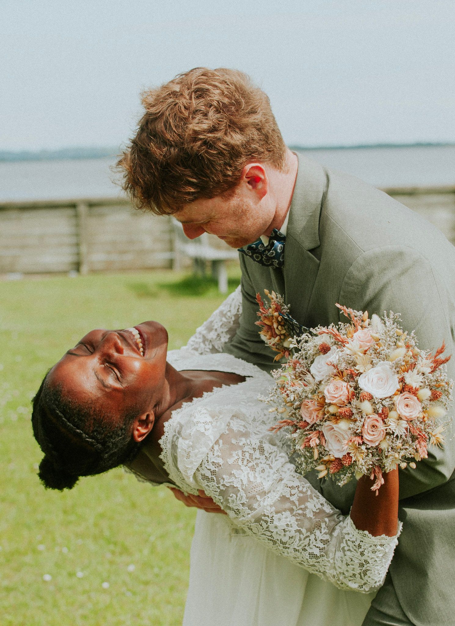Couple shares romantic dip pose with peach-colored bouquet in green field.