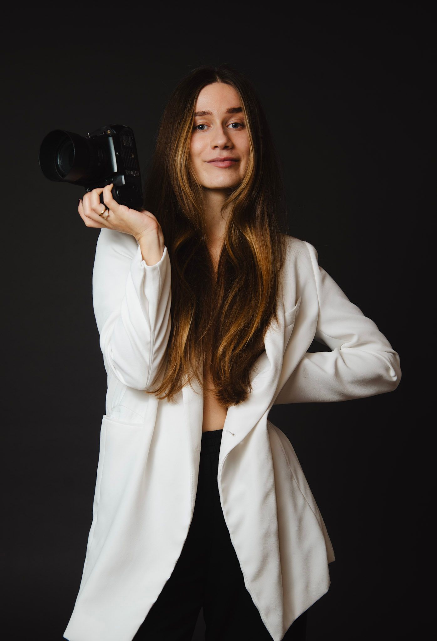 Professional photographer in white blazer poses with camera against dark background.
