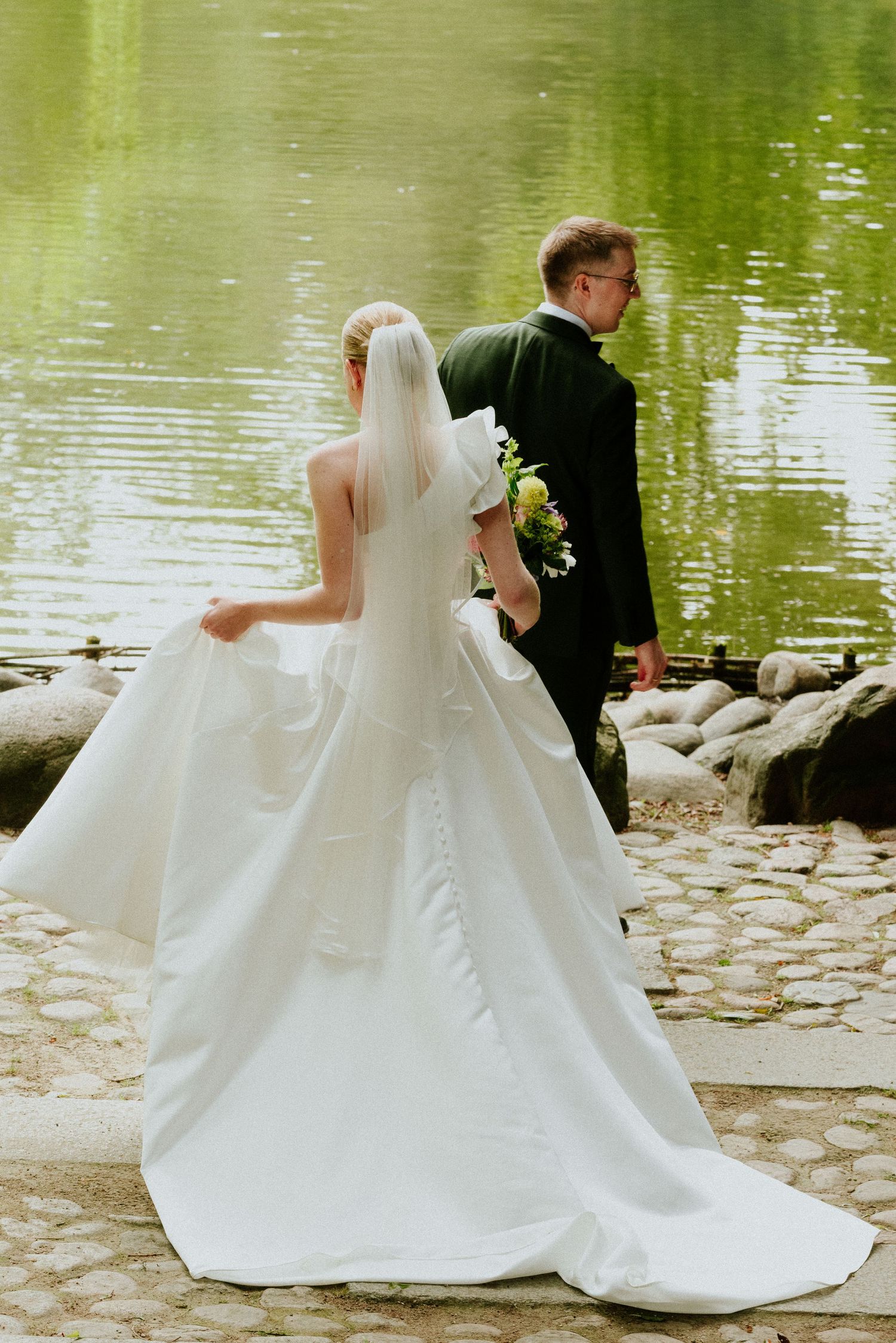 Newlyweds pose for photos by a serene pond with flowing white wedding dress.