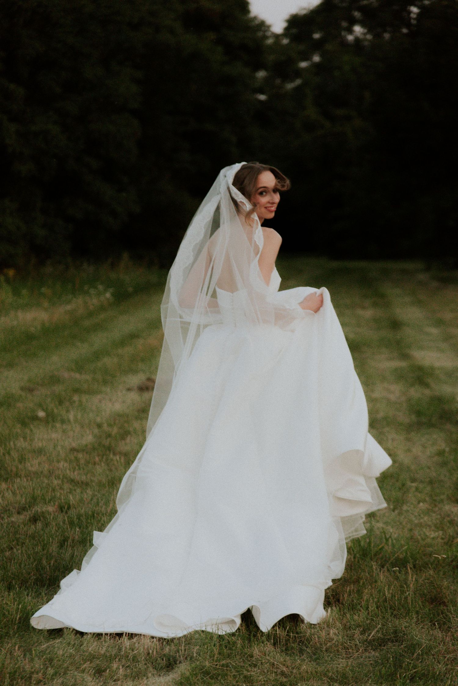 Bride in flowing white wedding gown twirls gracefully on grassy field at dusk.