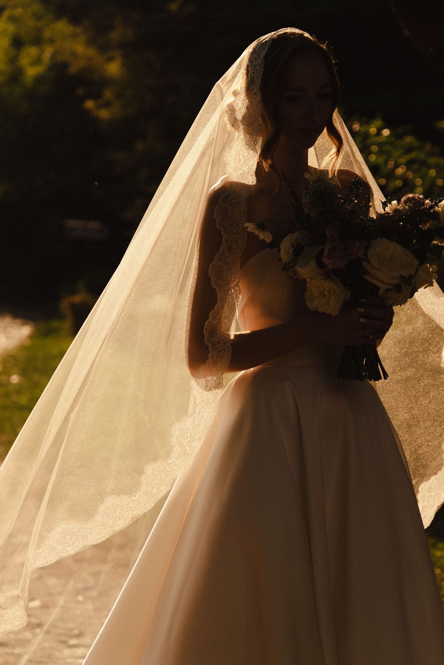 A bride in a flowing white wedding gown and veil holds a bouquet while backlit by warm golden sunlight.
