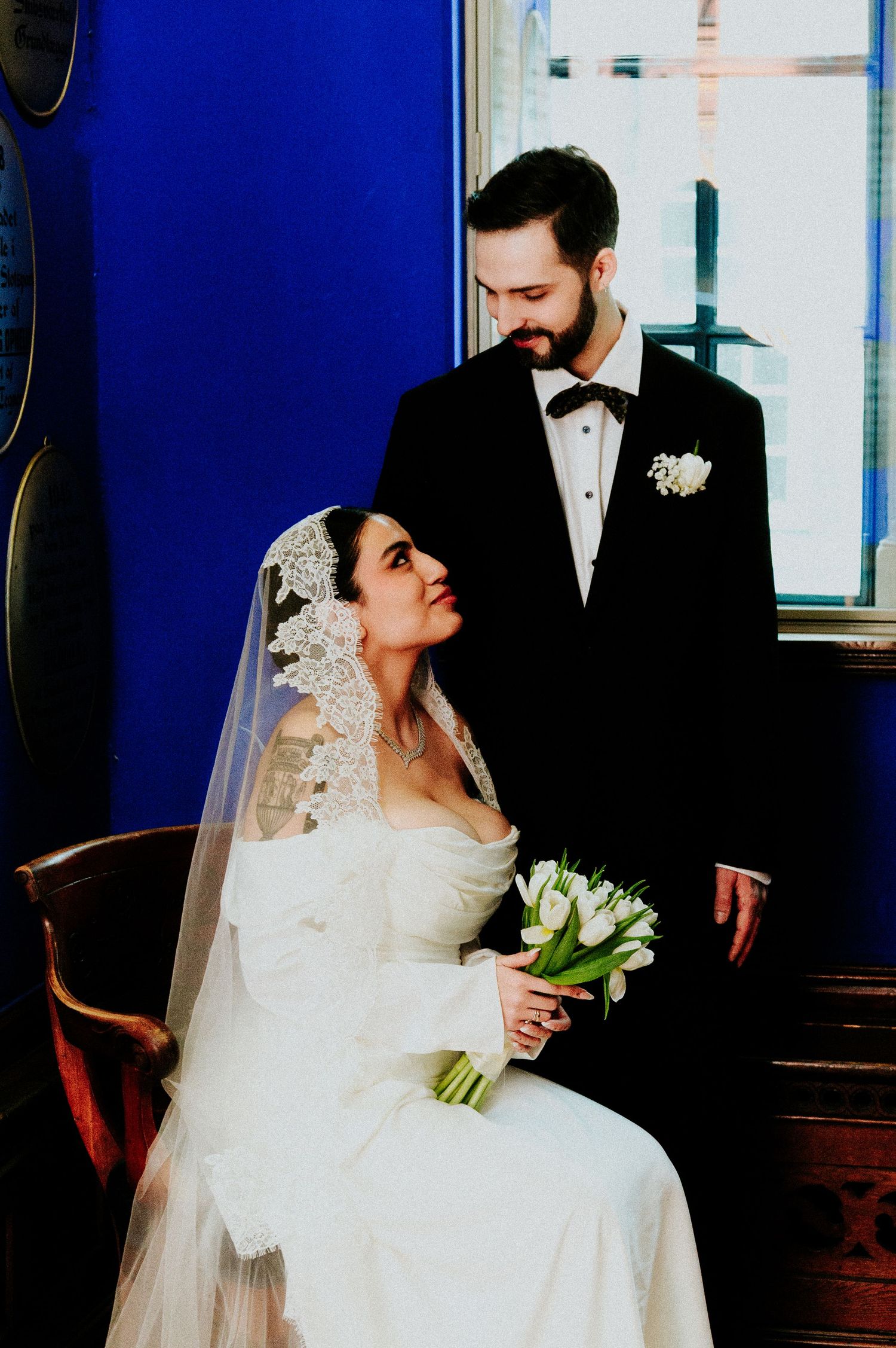 An elegant wedding portrait featuring a seated bride in white holding tulips beside her groom in a tuxedo.